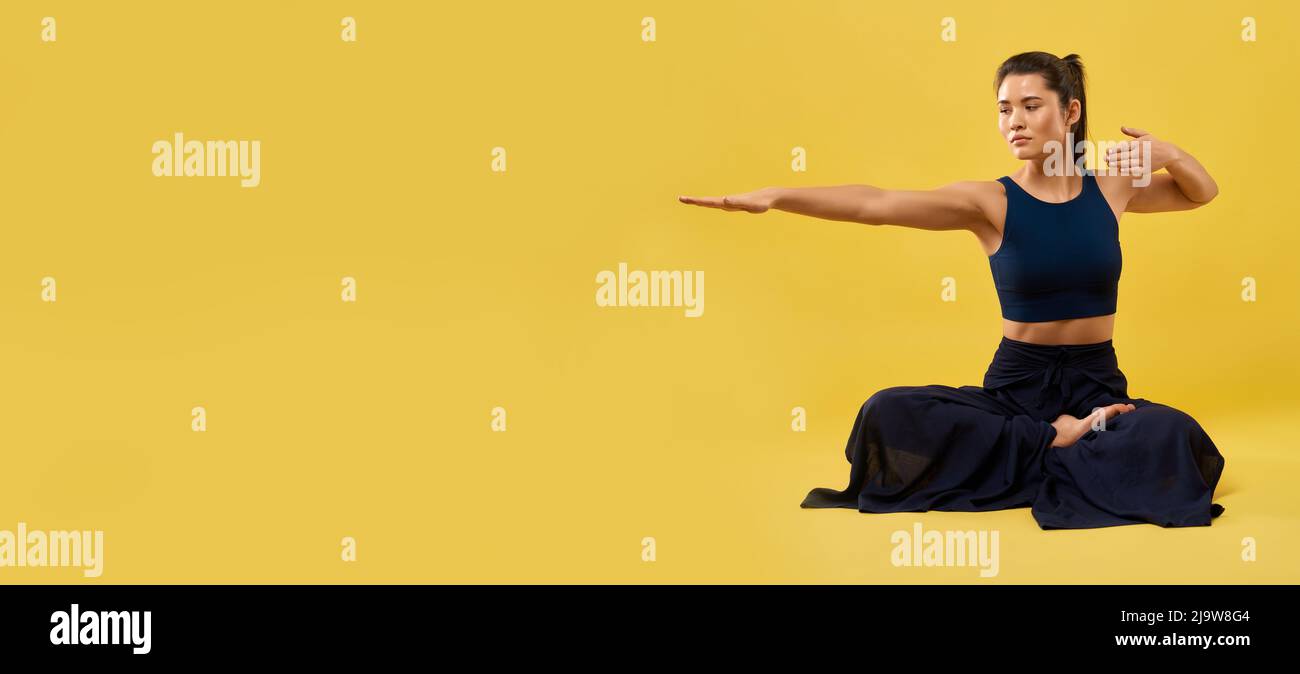 Bella ragazza che tiene il braccio destro allungato, mentre seduto in meditazione posa in interno. Vista frontale della signora che pratica yoga, mantenendo una mano piegata, isolato su sfondo arancione studio. Concetto di yoga. Foto Stock