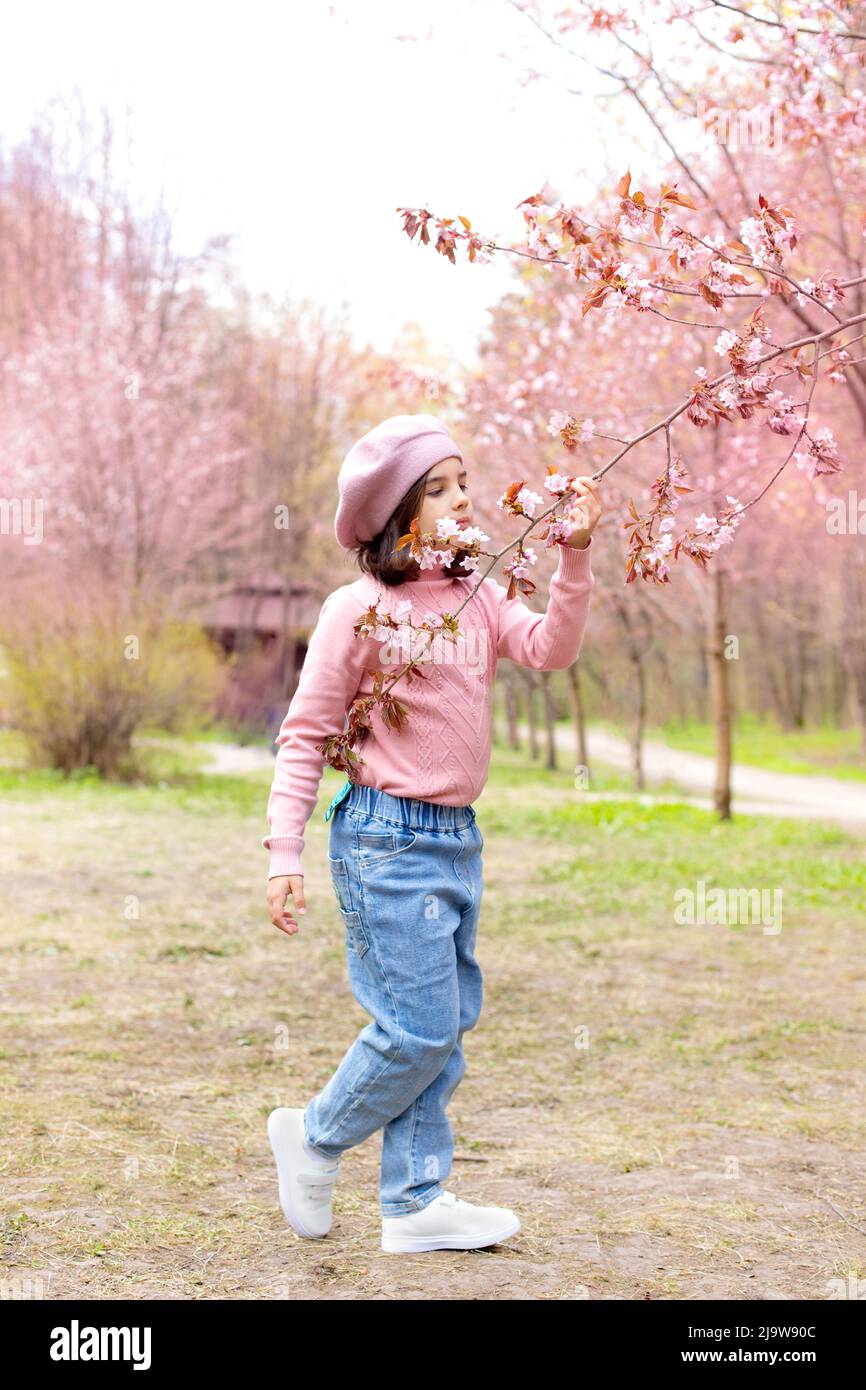 Una bambina in un ponticello rosa e jeans blu, annusa un ramo di fiori di ciliegia, nel parco durante il giorno Foto Stock
