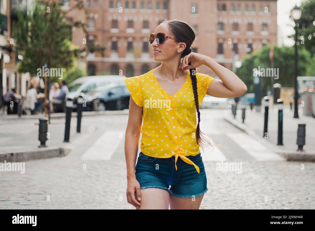 Ragazza in vacanza passeggiando per le strade di Madrid. Giorno di sole, con una t-shirt gialla e pantaloncini in denim. Occhiali da sole. Capelli in una lunga treccia. Copia s. Foto Stock