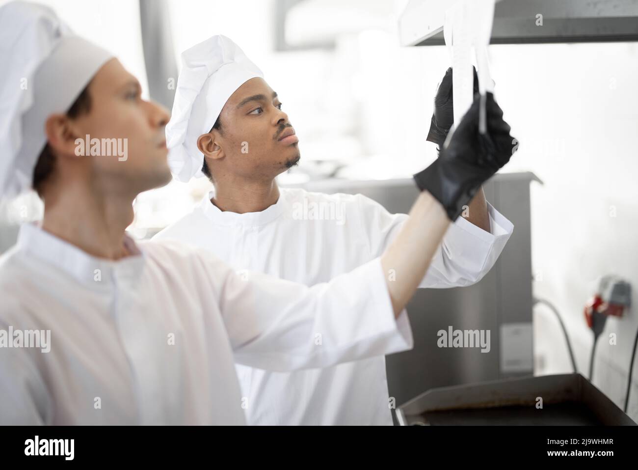 Due cuochi ben vestiti guardano gli assegni stampati con gli ordini mentre cucinano nella cucina professionale. Ragazzi latinoamericani e caucasici che lavorano insieme al ristorante Foto Stock