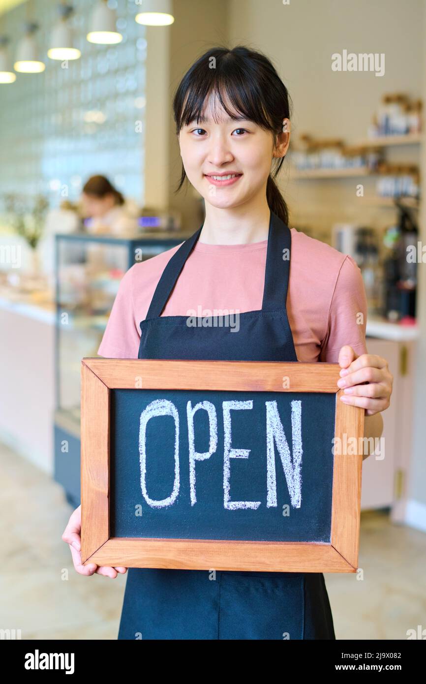 Ritratto della giovane cameriera asiatica in uniforme segno di tenuta e sorridendo alla macchina fotografica, lei apertura caffè Foto Stock
