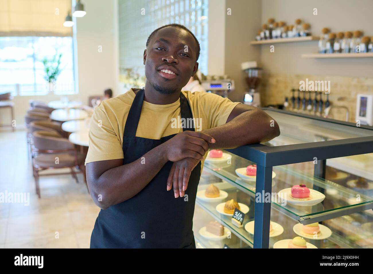 Ritratto del giovane proprietario africano in uniforme nera appoggiata sul bancone con cibo dolce e sorridente alla macchina fotografica durante il suo lavoro in caffetteria Foto Stock