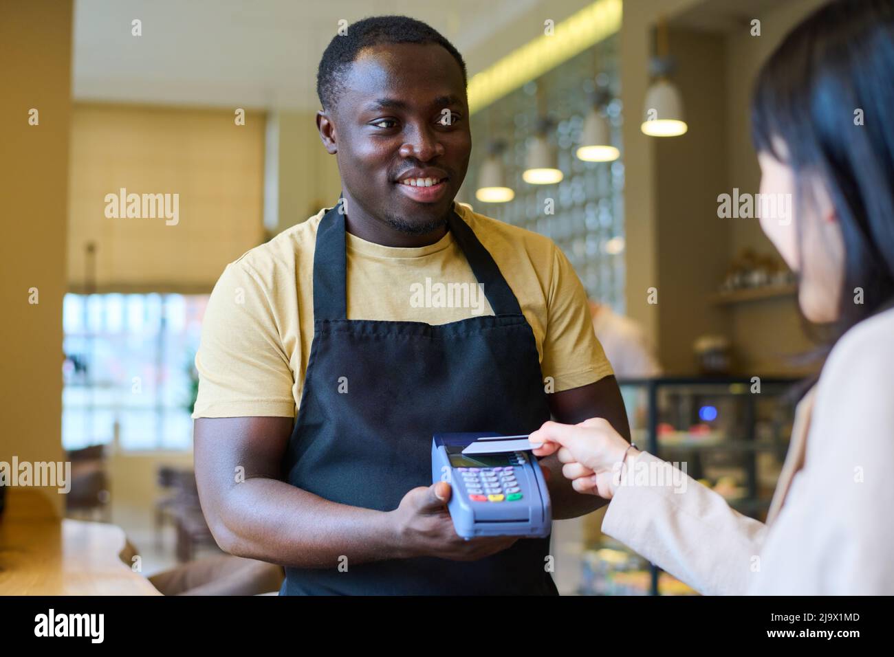 Giovane cameriere africano in uniforme tenendo terminale e sorridendo mentre il cliente paga con carta di credito dopo pranzo al ristorante Foto Stock