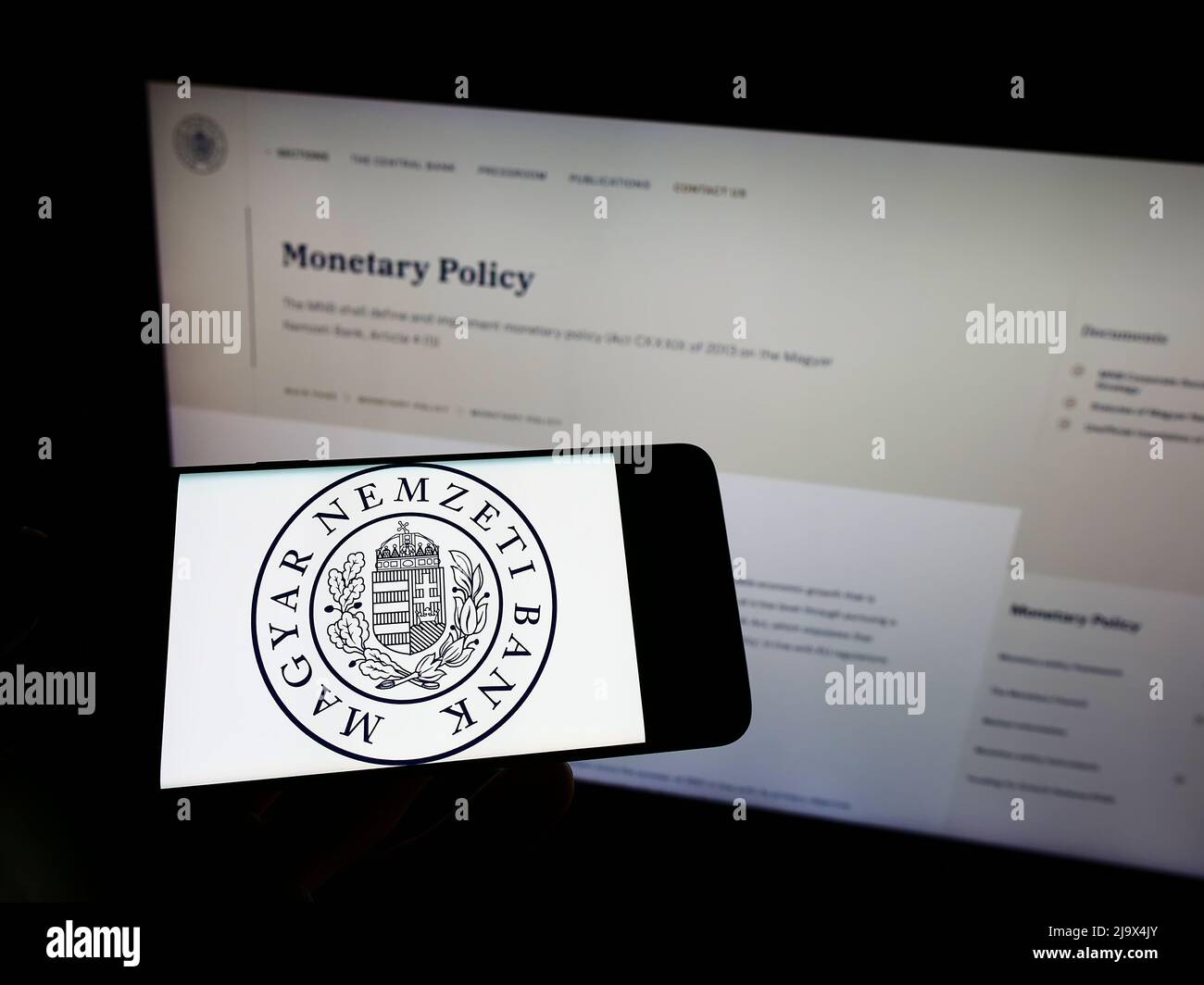 Persona che tiene il cellulare con il logo della Banca Nazionale Ungherese (Magyar Nemzeti Bank, MNB) sullo schermo di fronte alla pagina web. Mettere a fuoco sul display del telefono. Foto Stock