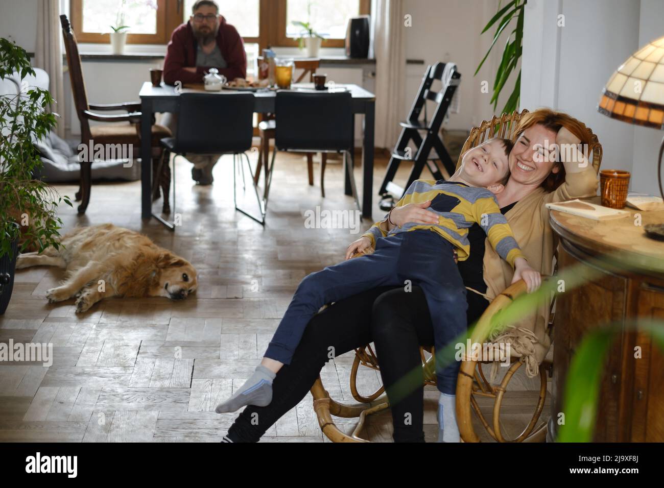 Madre con bambino e cane a casa su sedia a dondolo divertirsi insieme. Genitore con bambino rilassarsi su poltrona in casa d'epoca con animali domestici e. Foto Stock