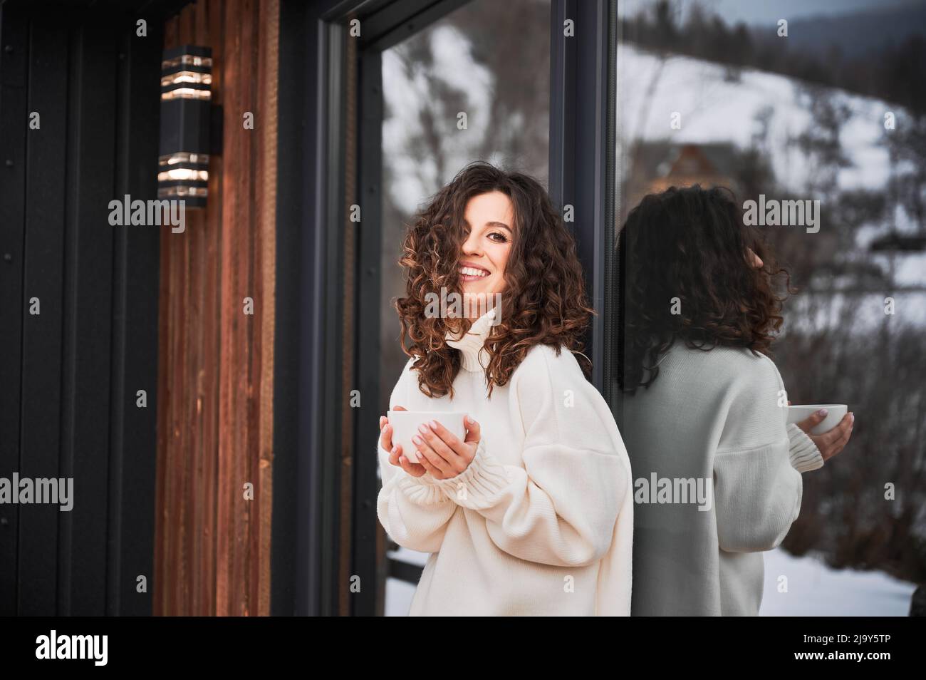 Giovane donna curly che riposa sulla terrazza della moderna casa di fienile in montagna. Ritratto di turista felice che tiene una tazza di tè, godendo in nuovo cottage in inverno. Foto Stock