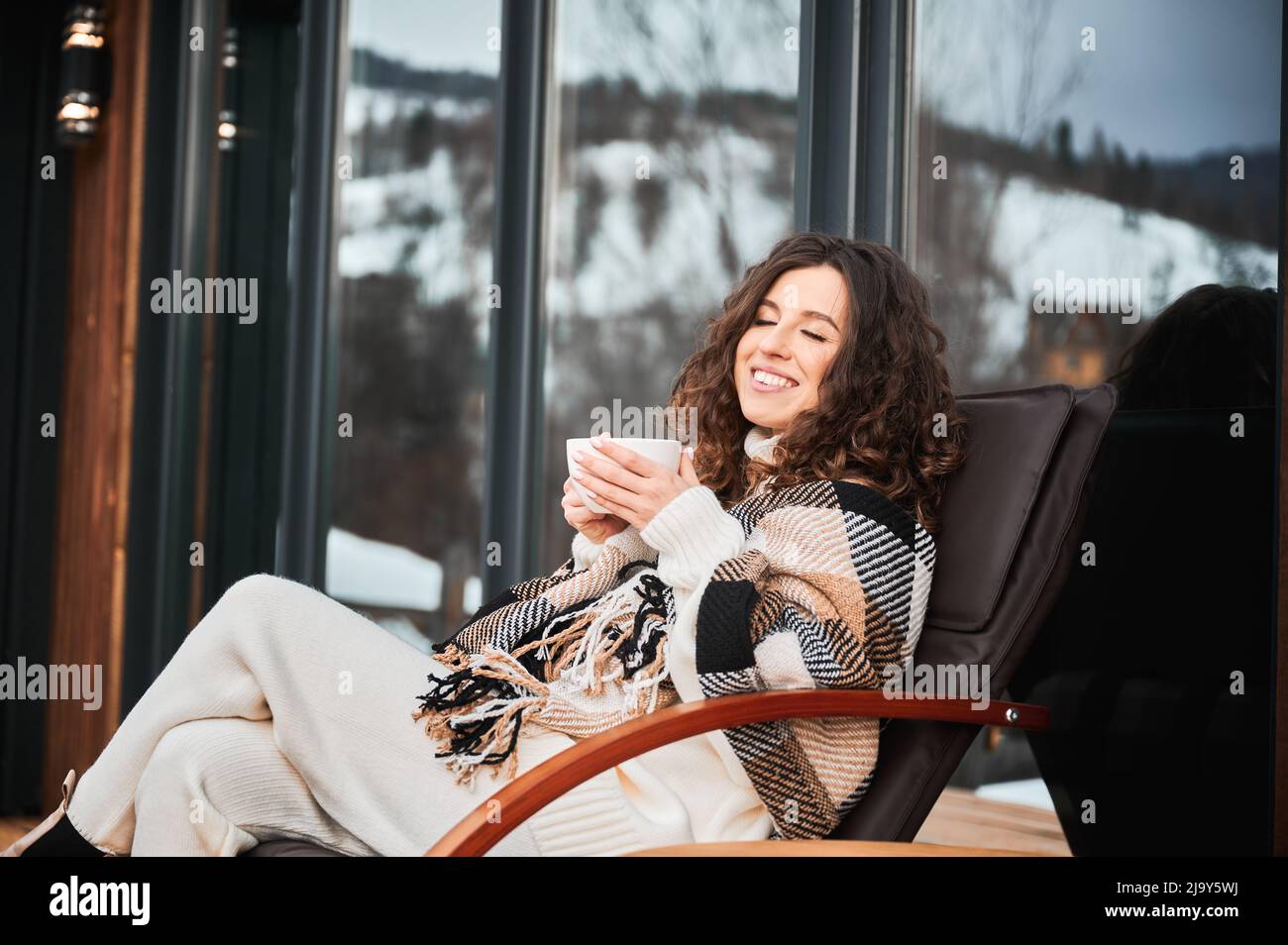 Giovane donna curly che riposa sulla terrazza della moderna casa di fienile in montagna. Felice turista femmina in plaid che tiene una tazza di tè, godendo in nuovo cottage in inverno. Foto Stock