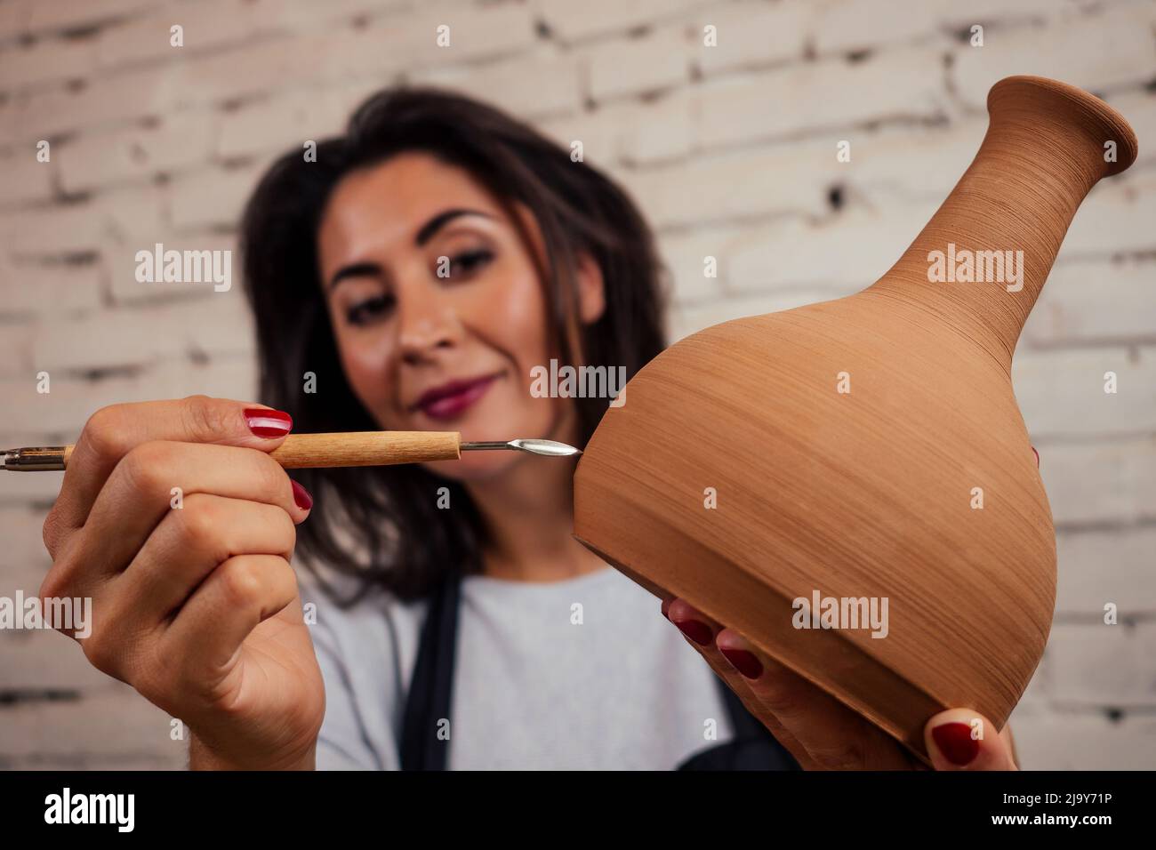 giovane e attraente vasaio femminile che lavora in uno studio con un vaso di argilla marrone nel laboratorio di artigianato tenendo l'attrezzo e la tazza in mano . business Foto Stock