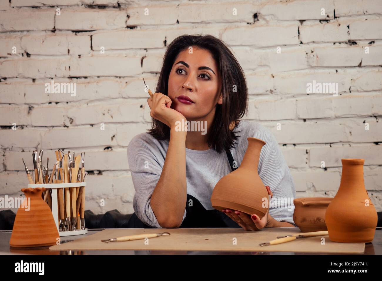 giovane e attraente femmina vasaio triste che lavora in uno studio con un vaso di argilla marrone nel laboratorio di artigianato tenendo l'attrezzo e la tazza in mano Foto Stock