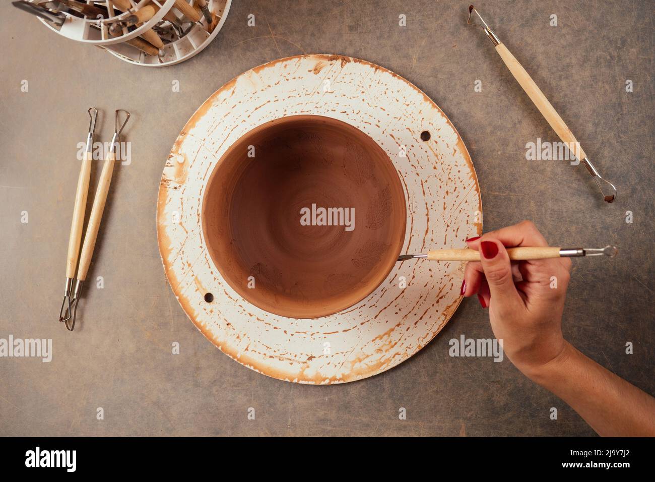 giovane e attraente vasaio femminile che lavora in uno studio con un vaso di argilla marrone nel laboratorio di artigianato tenendo l'attrezzo e la tazza in mano . business Foto Stock