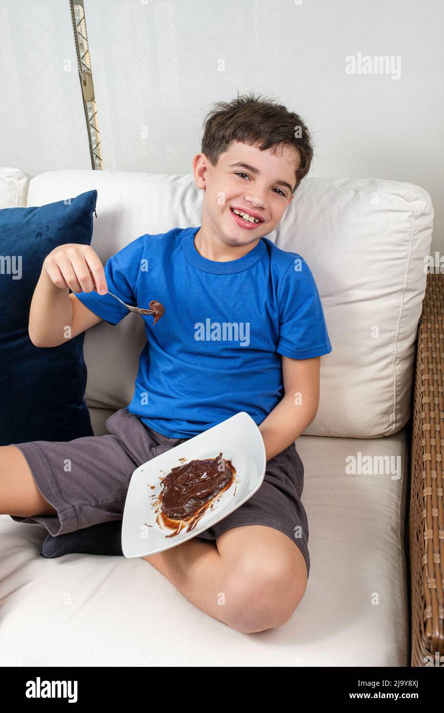 Sorridendo bambino di 8 anni seduto sul divano, tenendo un cucchiaio con dolce brigadeiro e guardando la macchina fotografica. Foto Stock