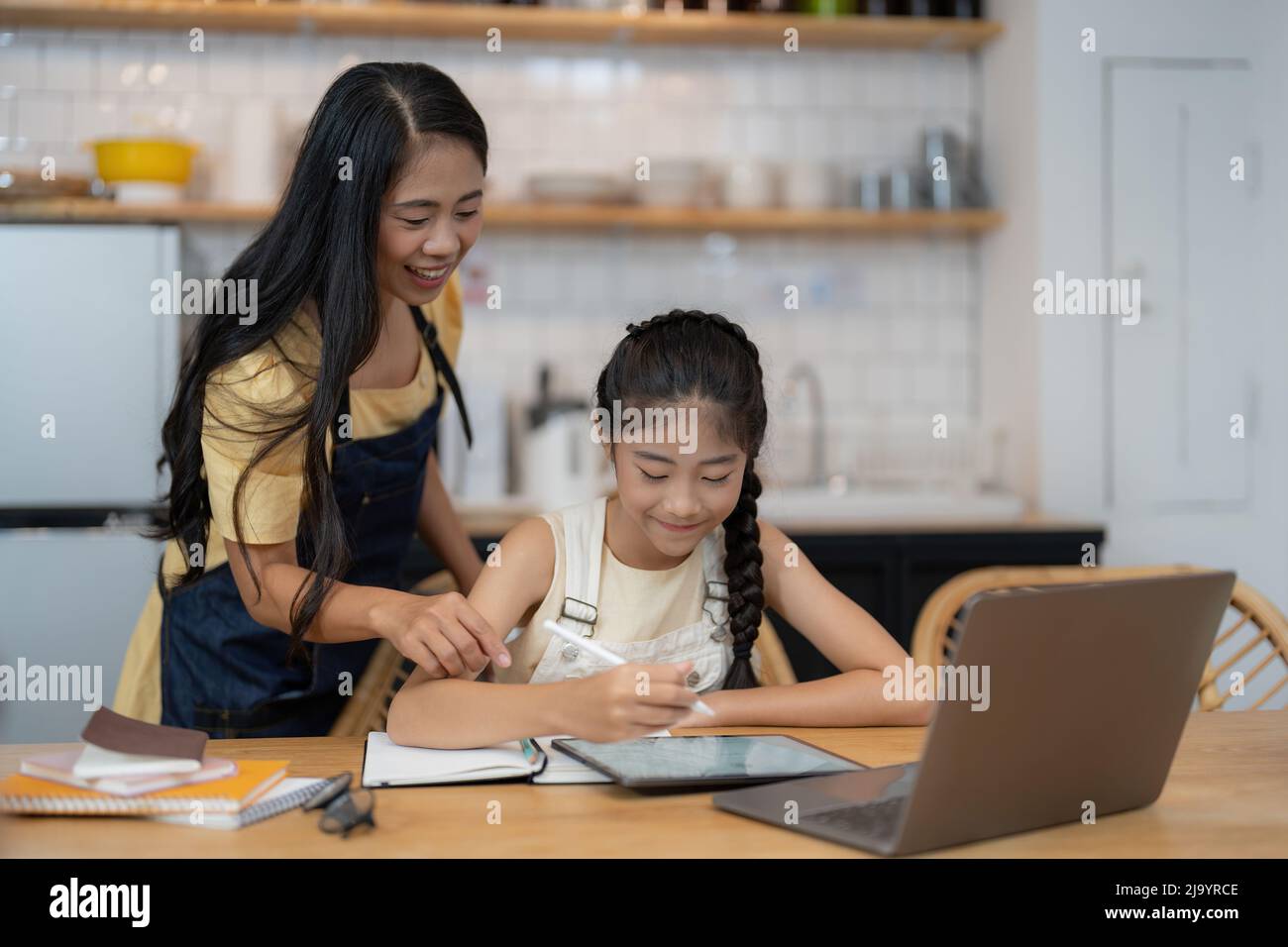 Madre asiatica e figlia piccola si siedono alla scrivania in cucina studiando in linea insieme, mamma biraciale e bambina piccola scrittura a mano del bambino, fanno i compiti, imparando Foto Stock