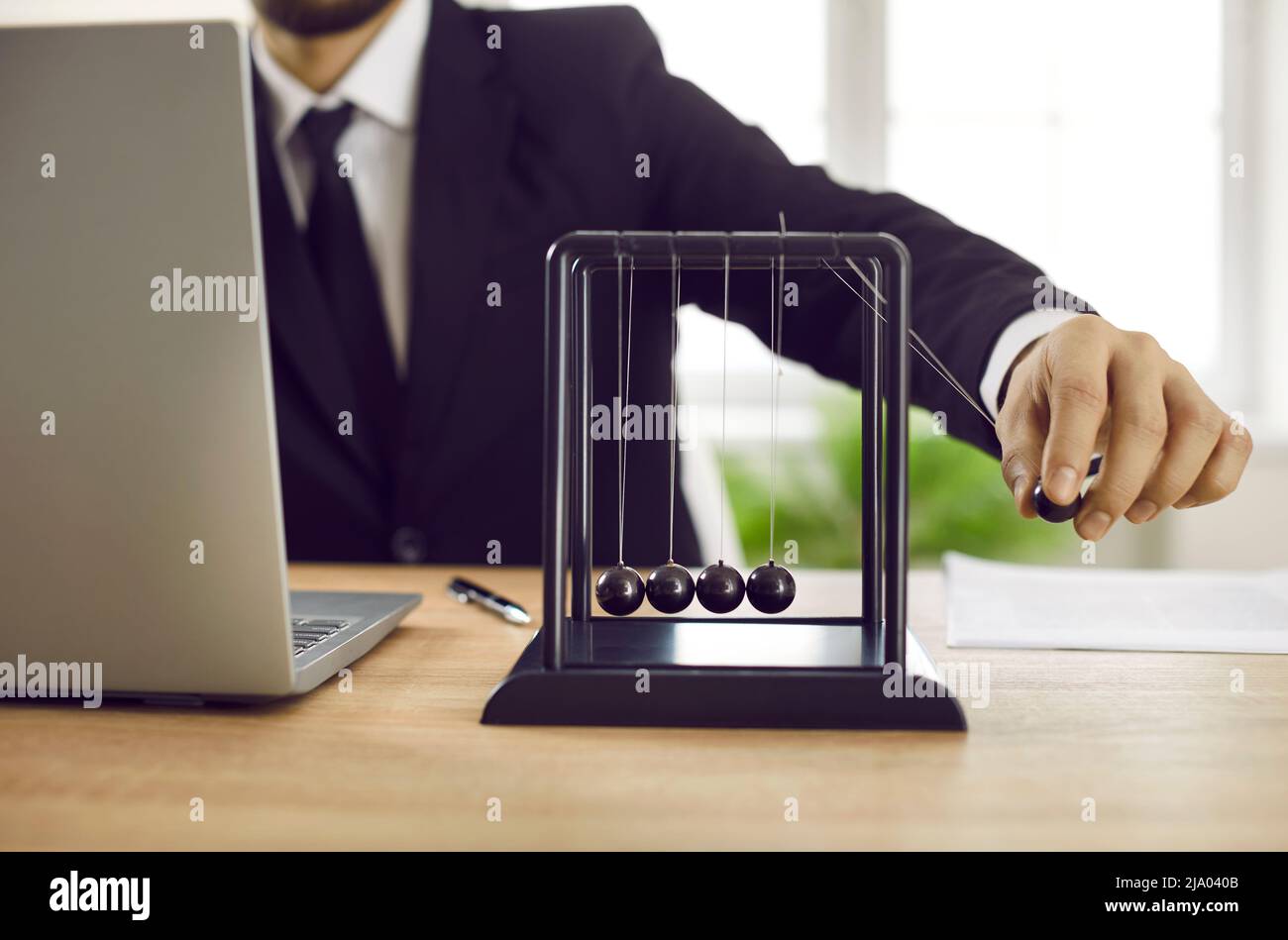 Uomo d'affari seduto alla scrivania d'ufficio con computer portatile e giocare con il pendolo di Newton Foto Stock
