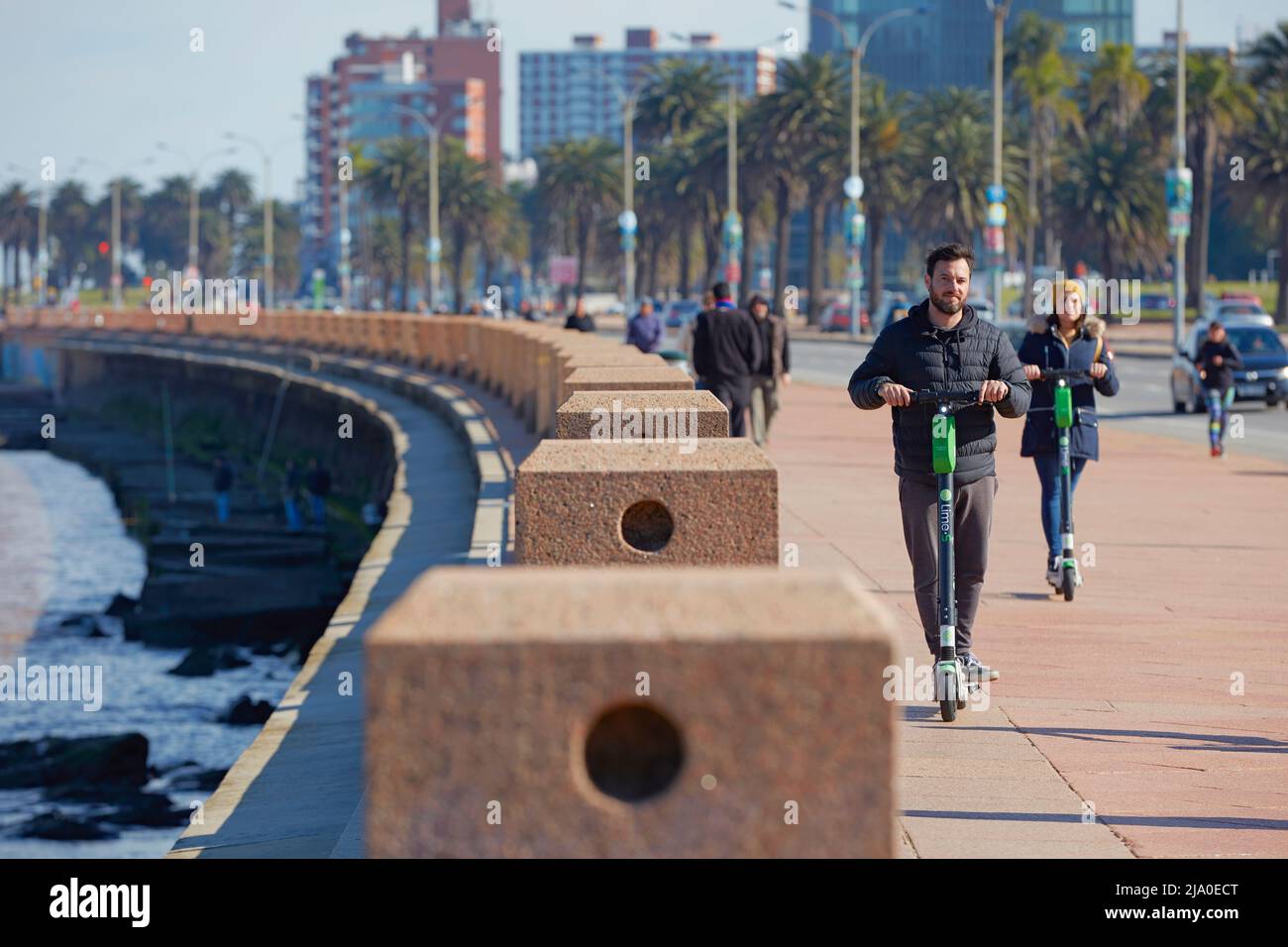 Due giovani cavalcano gli scooter elettrici lungo la Rambla di Montevideo, Uruguay. Foto Stock