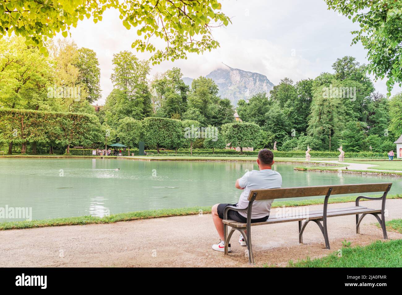 lone figura di uomo seduto su panca di fronte al laghetto con acqua turchese nel Parco Hellbrunn. parco in stile barocco. Stagione primavera. Salisburgo, Foto Stock