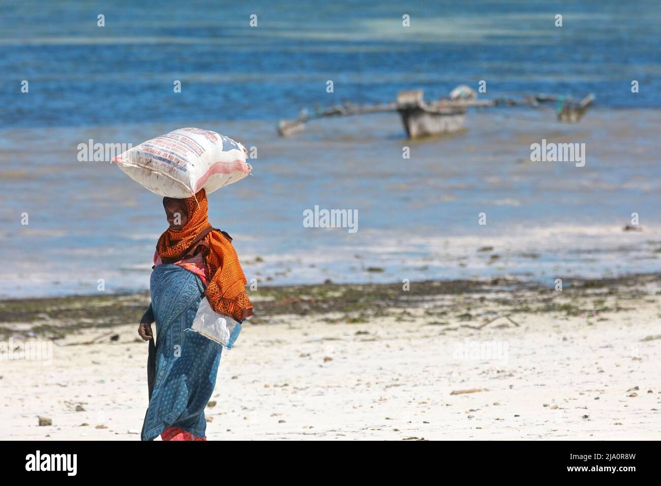 Una donna che porta un sacco di alghe sulla sua testa alla spiaggia di Jambiani con una tradizionale barca 'Ngalawa' sullo sfondo, Zanzibar, Tanzania, Africa. Foto Stock
