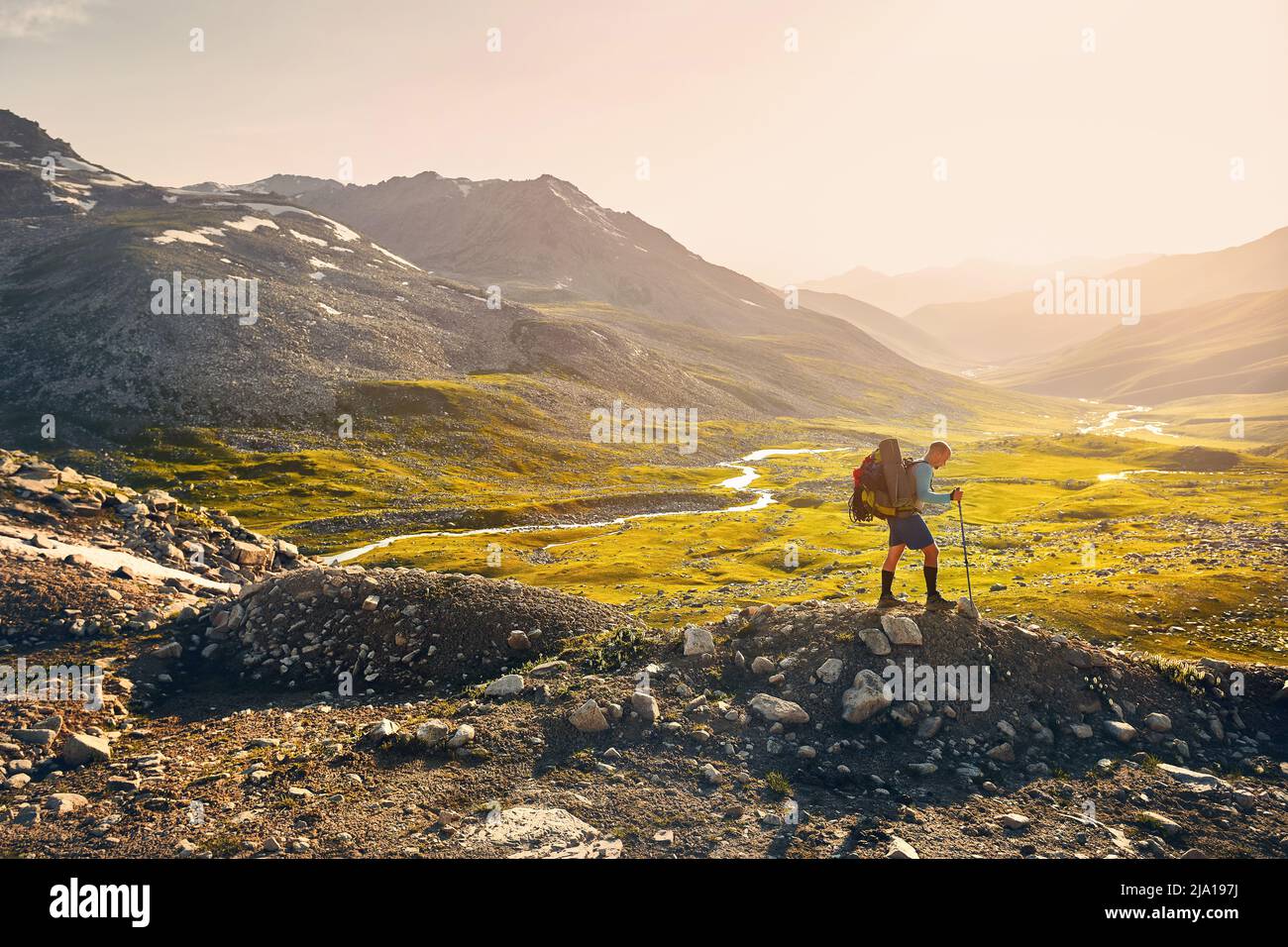 Uomo Hiker uomo turistico con grande zaino a piedi sulla roccia contro il tramonto e il fiume bello nella valle di montagna. Concetto all'aperto e trekking. Foto Stock