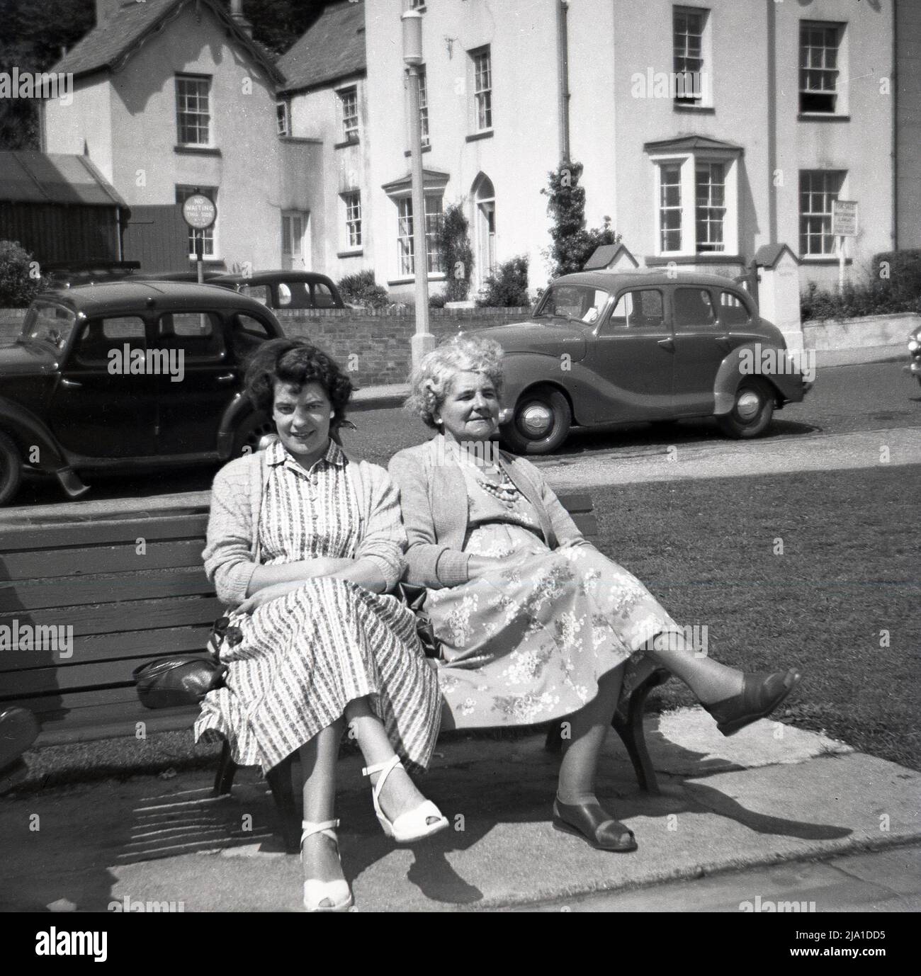 1950s, storica, una donna e sua madre che indossano gli abiti lunghi del giorno, seduti insieme su una panchina, prendendo l'aria di mare, Inghilterra, Regno Unito. Auto dell'epoca parcheggiate sul lato della strada. Foto Stock
