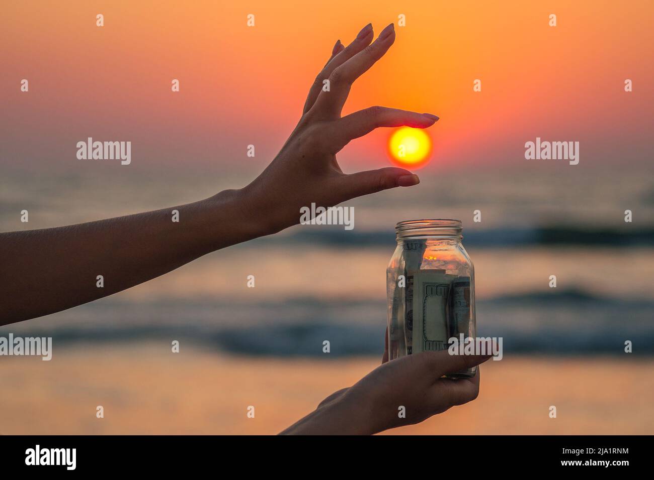 un vaso di vetro con monete e bollette sulla spiaggia contro il tramonto. il concetto di accumulo di denaro per i viaggi Foto Stock