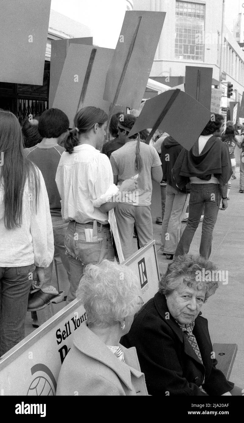 Due donne anziane aspettano ad una fermata dell'autobus mentre i giovani manifestanti che portano i cartelli passavano su Wilshire Boulevard a Los Angeles, CA. Foto Stock