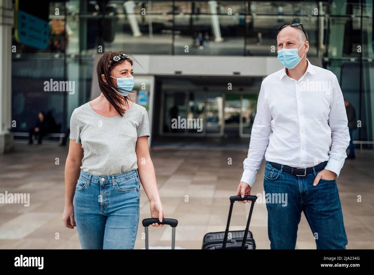 Un paio di amici in maschere protettive che chiacchierano mentre lasciano l'aeroporto con i loro bagagli Foto Stock