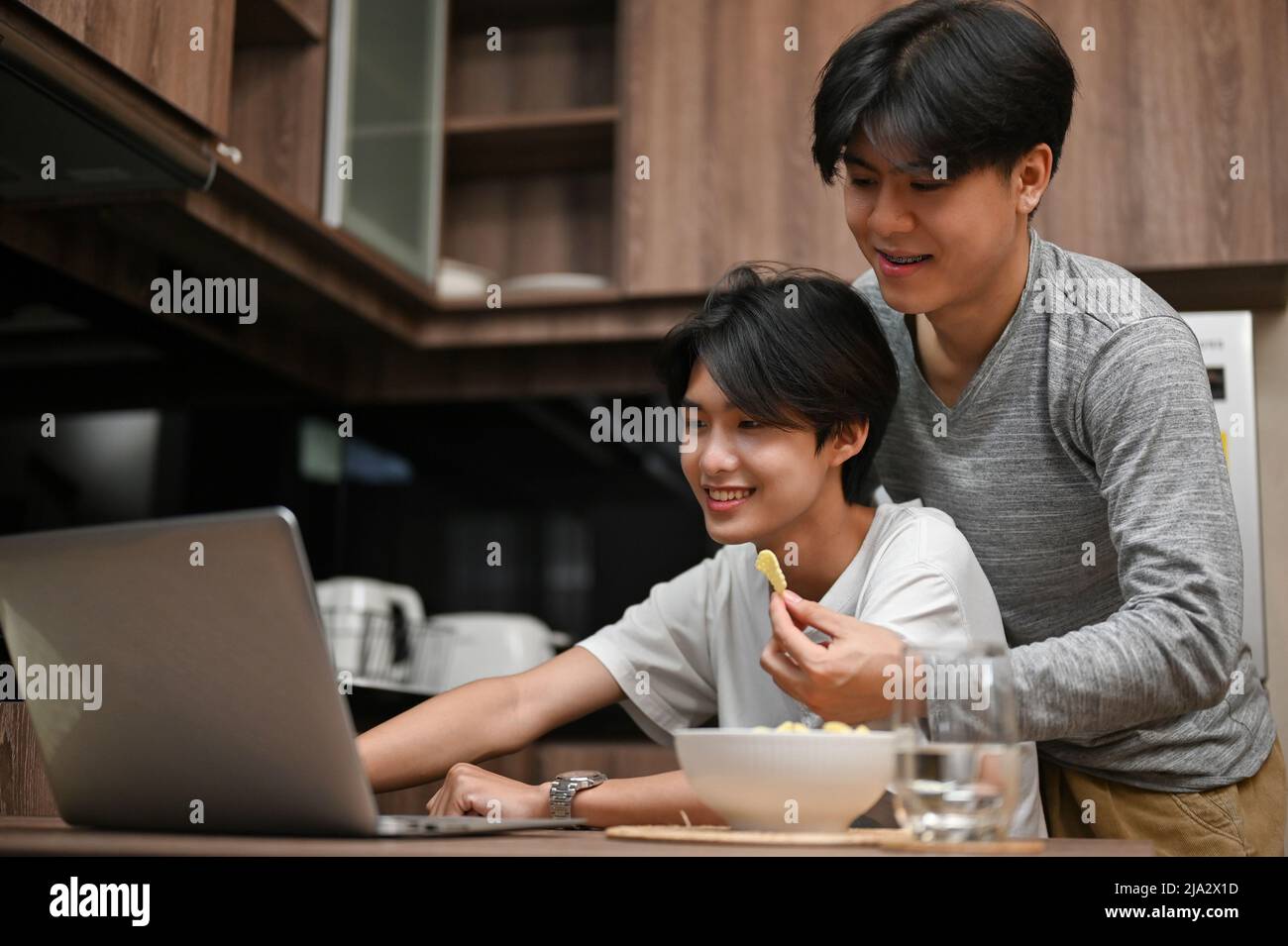 Un giovane gay asiatico attraente cerca di dare da mangiare uno spuntino al suo ragazzo mentre il suo ragazzo sta lavorando sul suo computer portatile in cucina. Foto Stock