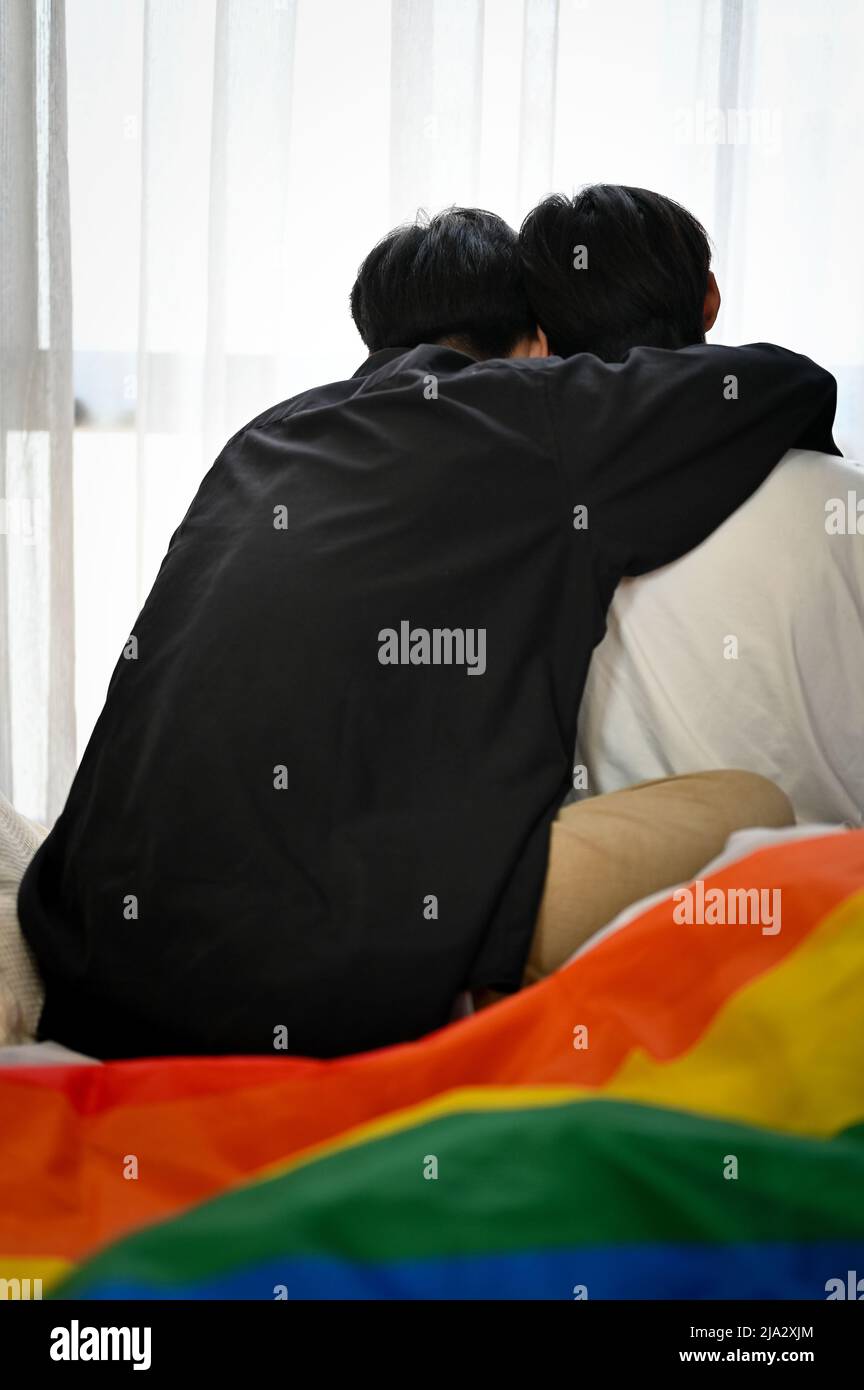 Back shot, Un'amorevole coppia di uomini gay asiatici adolescenti si siede insieme, si rilassa in salotto, mano intorno alla spalla del suo ragazzo. Coppia LGBT. Foto Stock