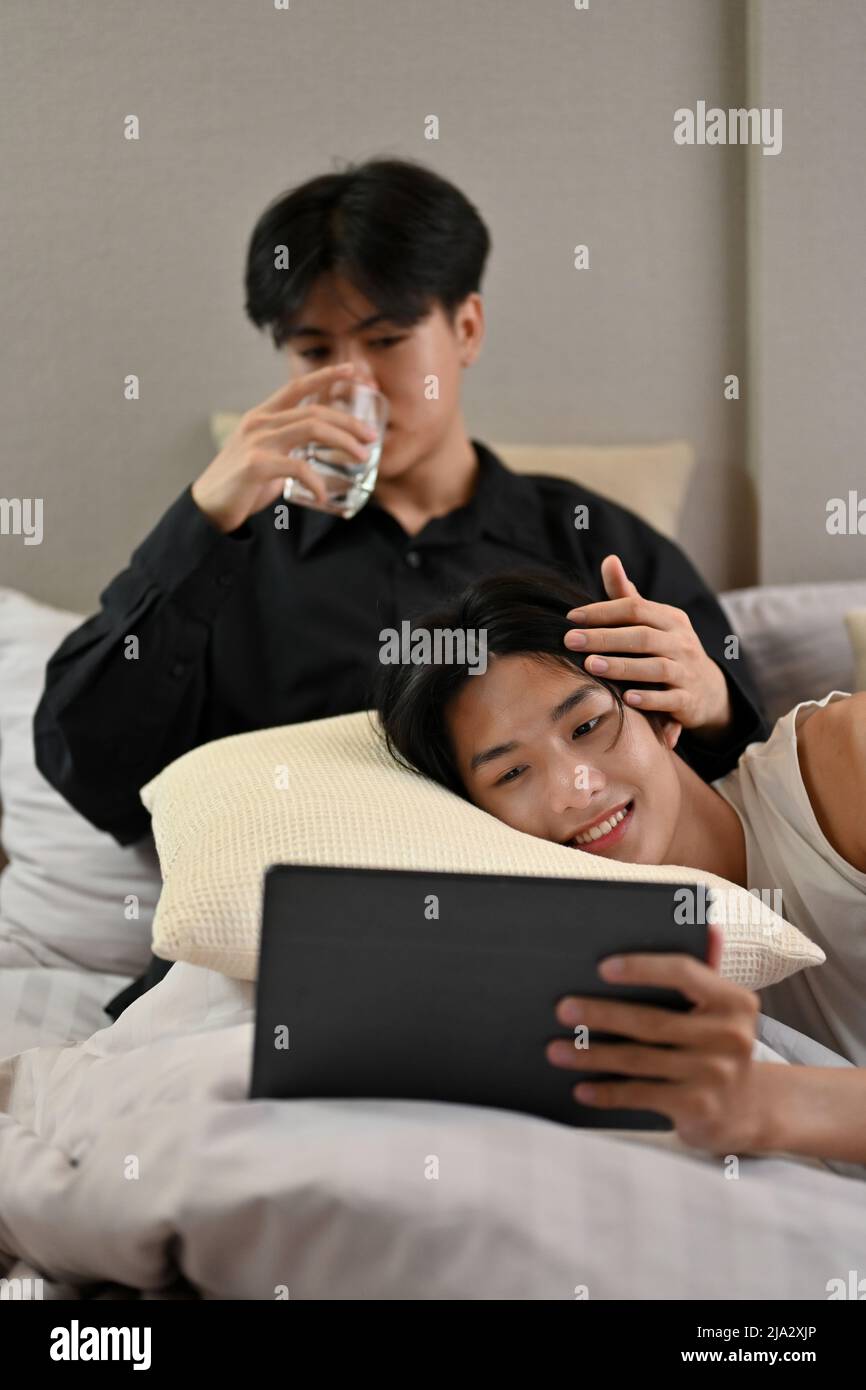 Bella coppia di giovani gay asiatici si rilassa in camera da letto, guardando un film su un tablet insieme. Un uomo che si trova sul cuscino sul suo giro ragazzo. Romanti Foto Stock