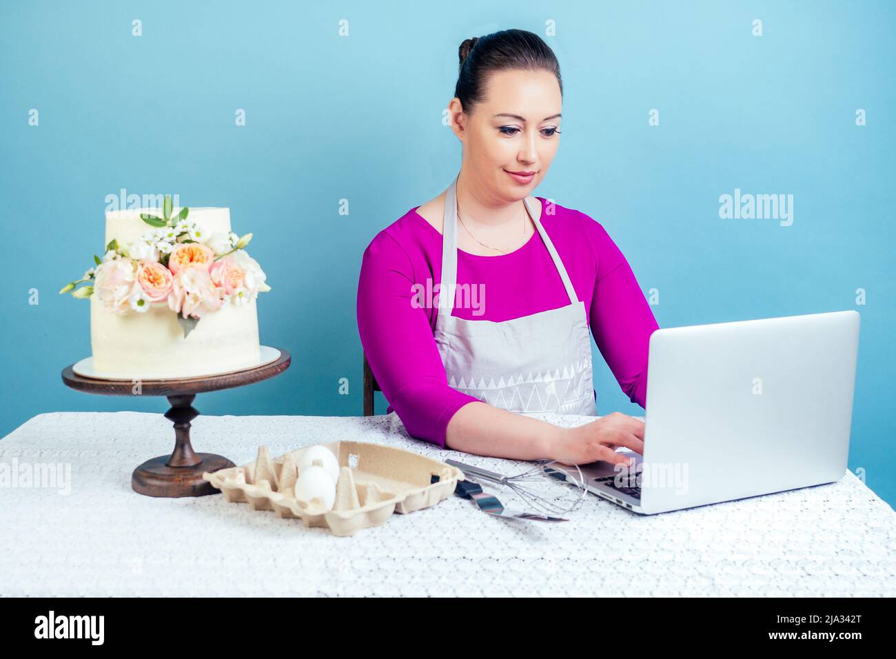 casalinga della pasticceria (pasticceria-cuoco) lavora con un computer portatile vicino alla torta bianca di nozze a due livelli con fiori freschi su un tavolo con una tovaglia di pizzo in Foto Stock