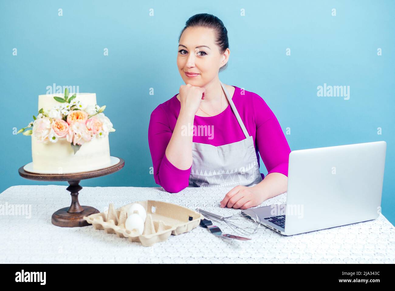casalinga dolciaria (pasticciere-cuoco) donna d'affari lavora con un computer portatile accanto alla torta di nozze bianca a due livelli con fiori freschi su un tavolo con un merletto Foto Stock