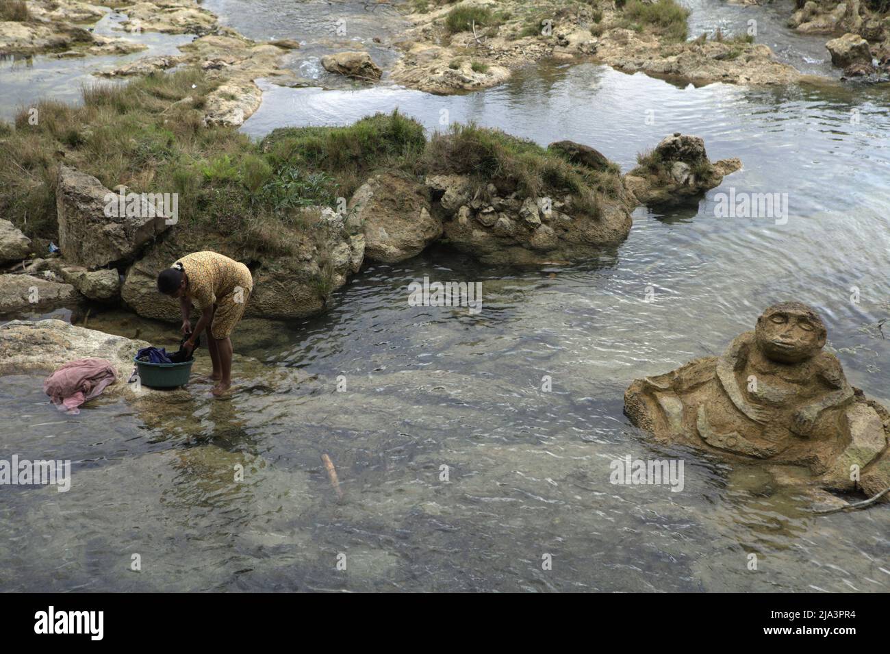 Una donna che lava abiti su un ruscello vicino a una pietra scolpita di figura umana vicino a Waikelo Sawah, una fonte d'acqua rara a Sumba, un'isola regolarmente colpita dalla siccità, che si trova nel villaggio di Tema Tana, East Wewewa, Southwest Sumba, East Nusa Tenggara, Indonesia. Foto Stock