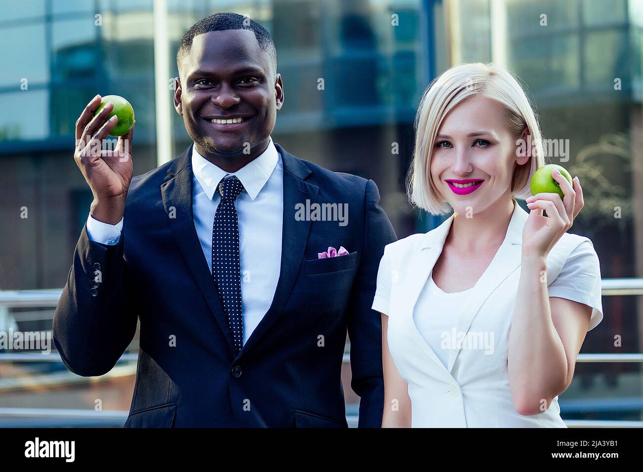 Bell'uomo d'affari Afro in un abito elegante nero con attraente signora bionda business mangiare una mela verde sullo sfondo strada grattacielo ufficio Foto Stock