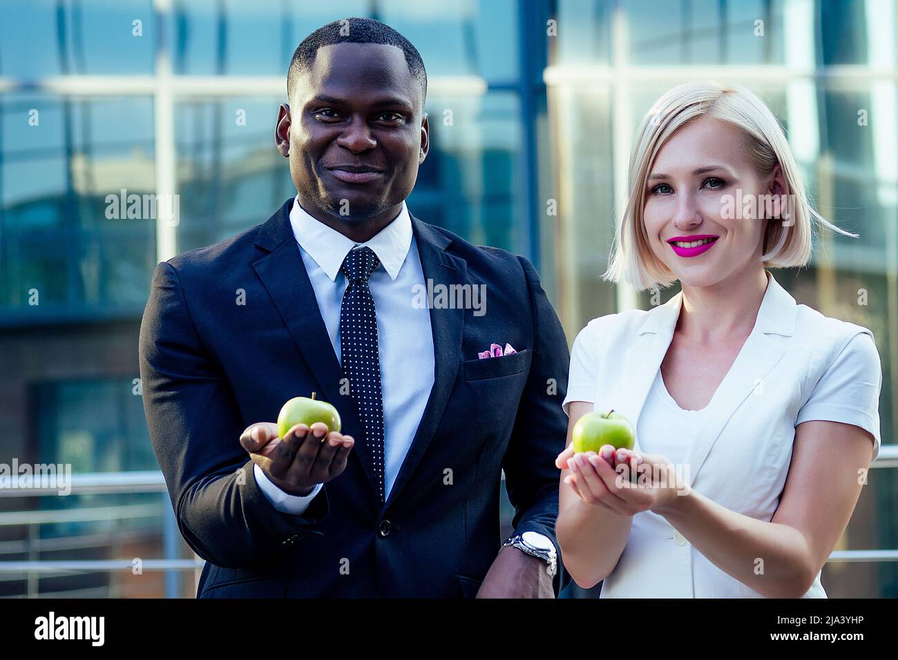 Bell'uomo d'affari Afro in un abito elegante nero con attraente signora bionda business mangiare una mela verde sullo sfondo strada grattacielo ufficio Foto Stock