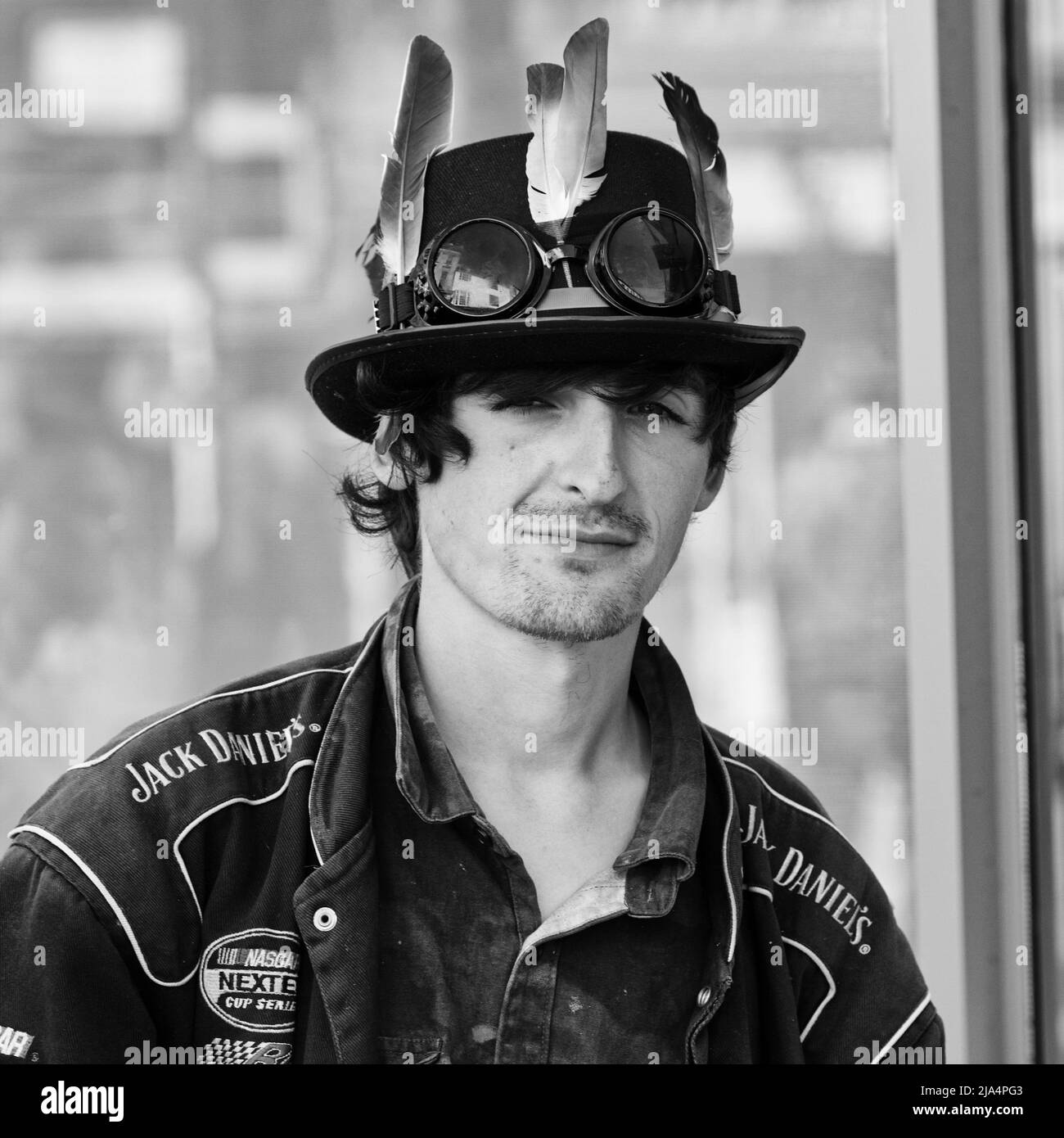 Abbigliamento culturale da banco su questo giovane maschio bianco che ha posto per questo ritratto di strada indossando una giacca Jack Daniels e un cappello con top in piuma Foto Stock