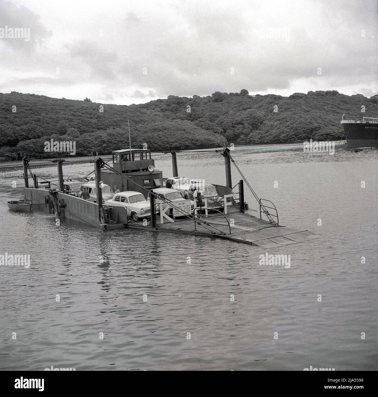 1950s, storico, il traghetto del re Harry, trasportante le automobili dell'epoca e della gente, attraverso le strade di Carrick raggiungono l'estuario del fiume Fal in Cornovaglia, Inghilterra, Regno Unito. Nel 1888 fu istituito un piccolo ponte di traghetti a catena o a fune che utilizzò un motore a vapore per tirare le catene e questo fu utilizzato fino all'introduzione di un motore a bisello nel 1956. Ancora in funzione oggi, il fiume Fal traversata è uno dei più scenografici viaggi in traghetto del mondo e una delle cinque catene ferrys ancora in funzione in Inghilterra. A destra della foto, la nave, British Knight, una nave petroliera, si stese sul fiume. Foto Stock