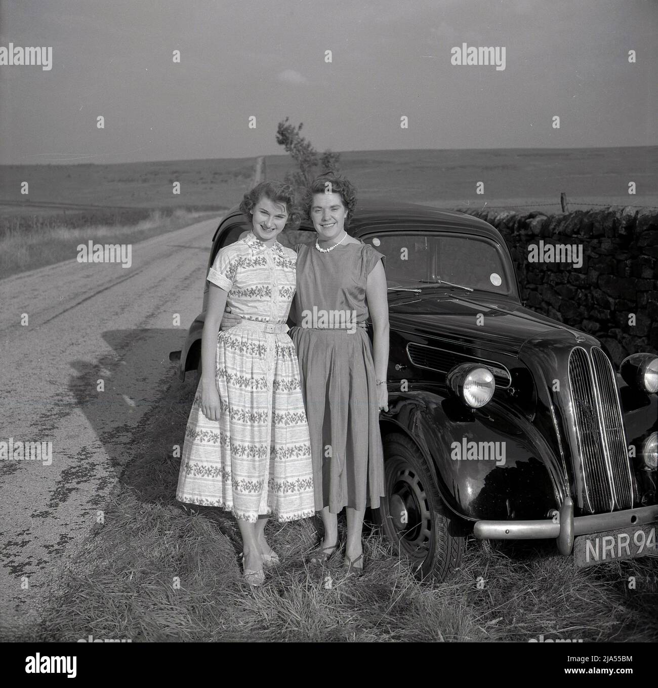 1950s, storica, due Signore, forse madre e figlia, nei lunghi abiti dell'epoca, in piedi per la loro foto con la loro auto Ford Anglia, che è parcheggiata su una verga di erba accanto a un muro di pietra su una strada di campagna, Inghilterra, Regno Unito. Foto Stock