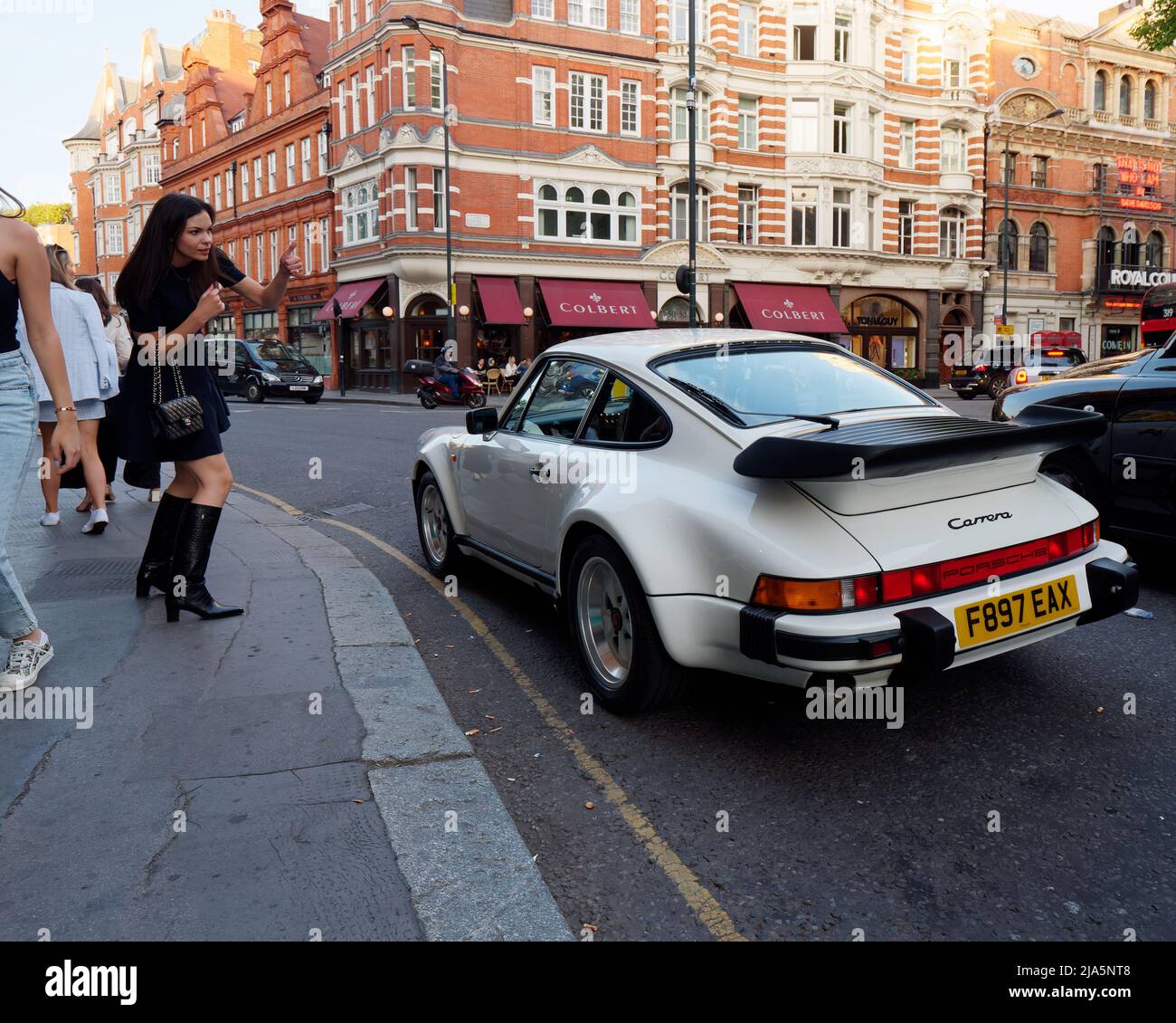 Elegante donna in tutto nero con gonna corta e scarponi segnali per un taxi, con una Porsche 911 bianco auto sportiva. Sloane Square, Chelsea, Londra. Foto Stock