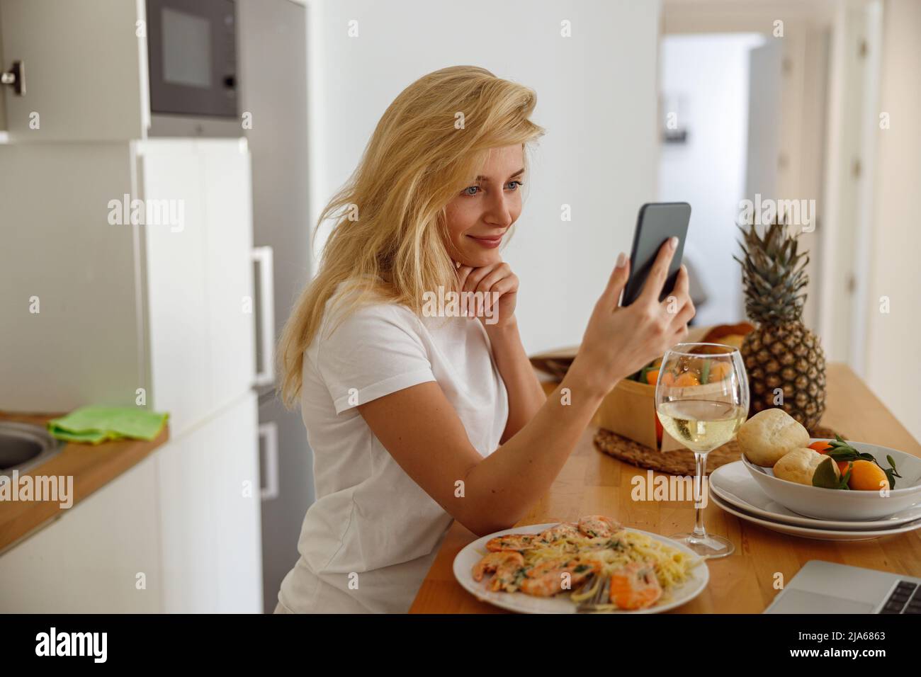 Ritratto della giovane bionda caucasica che tiene smartphone in cucina durante la cena. Foto Stock