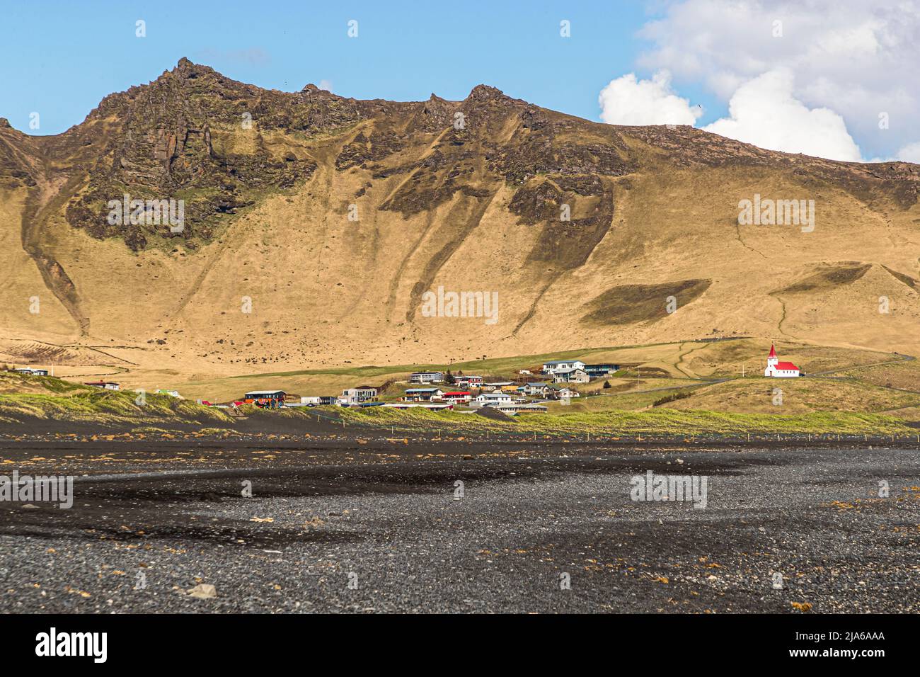 Il villaggio di Vík í Mýrdal si trova qui sotto una montagna (forse Reynisfjall) sulla costa meridionale dell'Islanda; la chiesa Víkurkirkja dai tetti rossi su una collina serve alla comunità come rifugio di emergenza in caso di eruzione del vulcano Katla, le cui inondazioni glaciali potrebbero allagare l'insediamento nel caso peggiore. La chiesa è stata consacrata nel 1934. In primo piano, la pianura di sabbia nera si estende verso il villaggio. Foto Stock