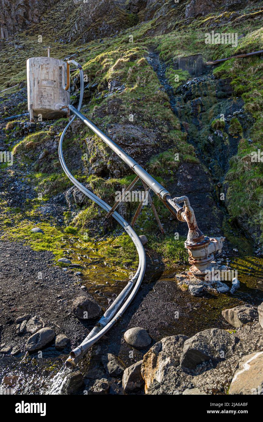 La piscina di Seljavallalaug è riscaldata qui tramite un tubo di metallo corroso con una valvola che convoglia l'acqua termale riscaldata geotermicamente lungo il pendio; dal giunto del tubo sale vapore. Costruita nel 1923 ai piedi del vulcano Eyjafjallajökull, la piscina di 25 per 10 metri è tra le più antiche piscine sopravvissute in Islanda. Foto Stock