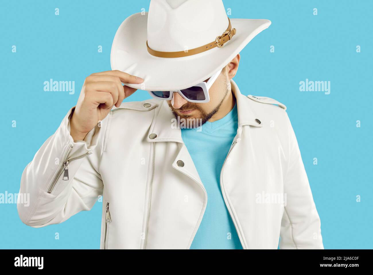 Uomo elegante e sicuro con espressione misteriosa che tiene il cappello da cowboy su sfondo blu chiaro. Foto Stock