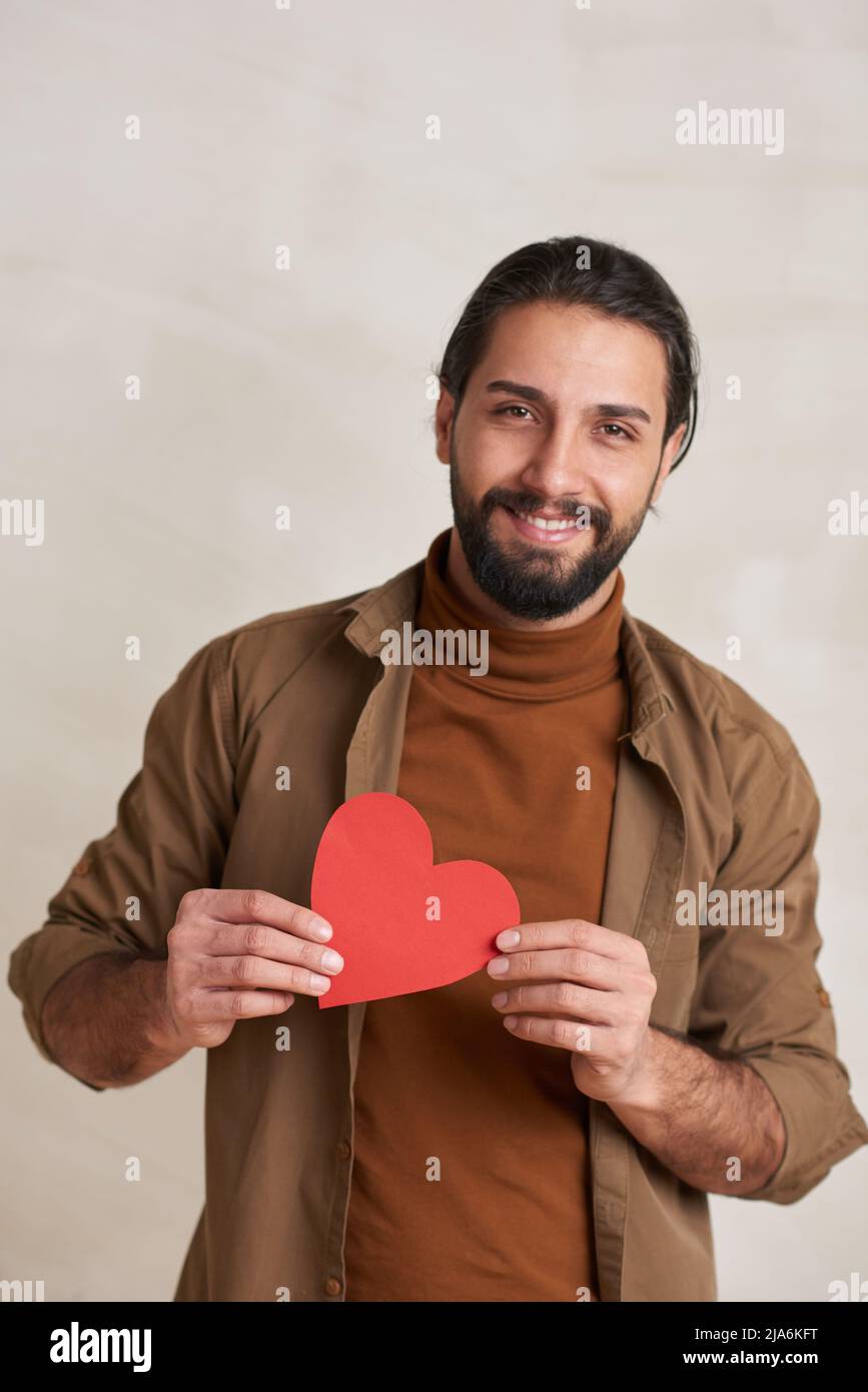 Verticale vita in alto ritratto di bel borsato giovane uomo che tiene rosso carta cuore sorridente alla fotocamera, copia spazio Foto Stock