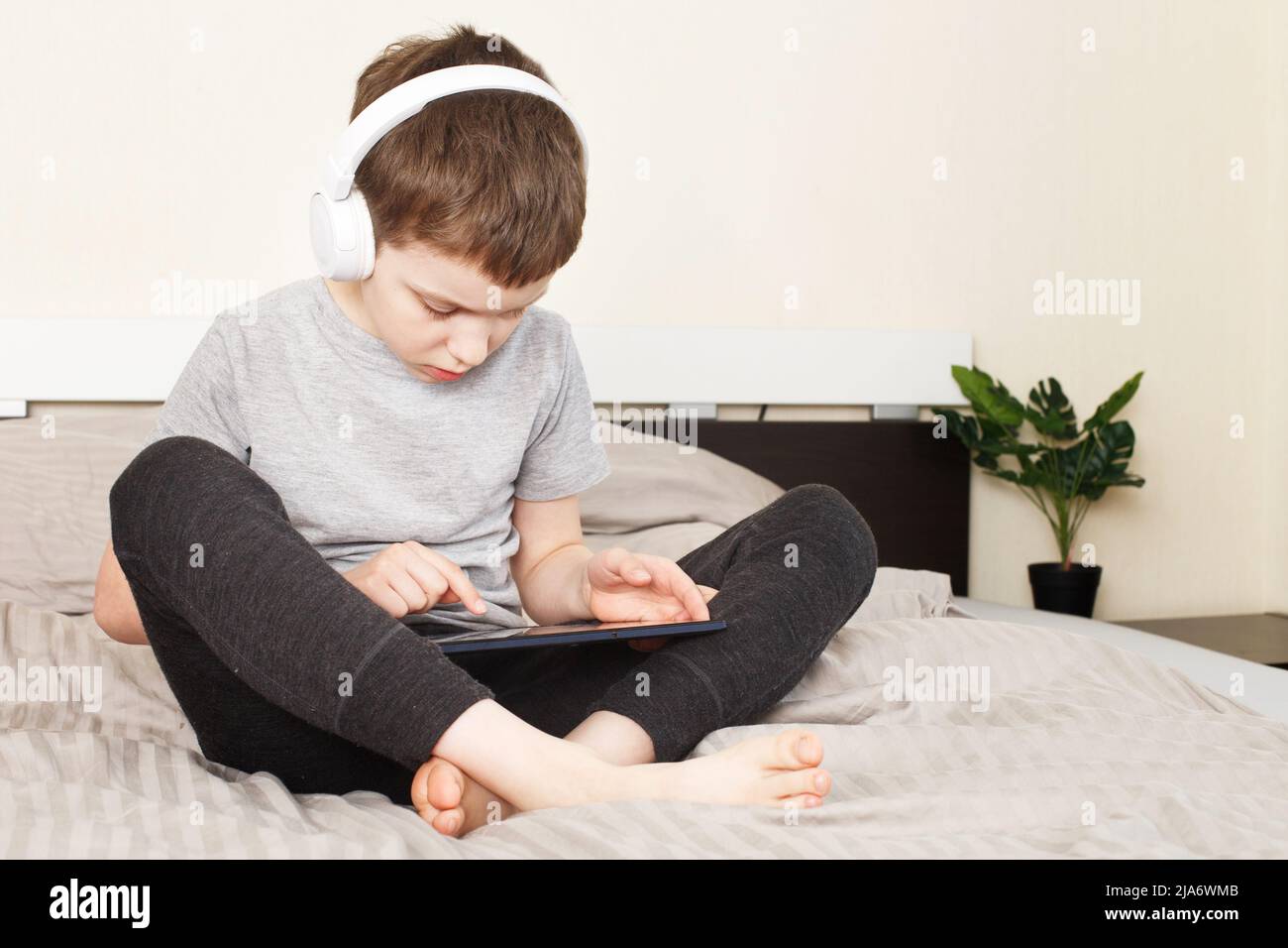 bambino seduto a letto a casa con tablet pc e lettura online, apprendimento o ascolto di musica con cuffie wireless Foto Stock