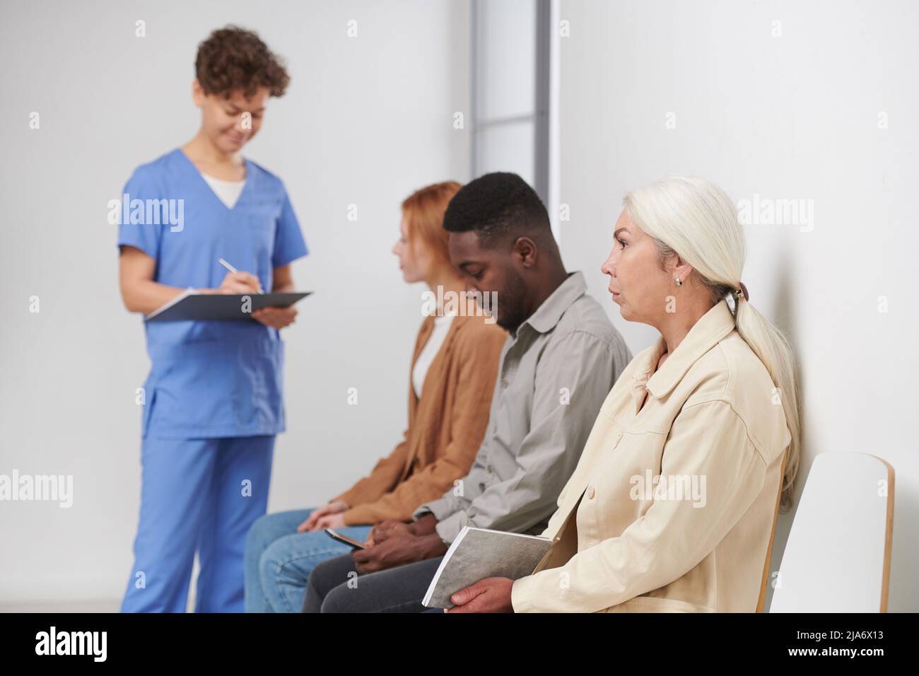Gruppo di tre persone sedute in coda in attesa di consultazione in ospedale moderno, operatore medico in piedi vicino a loro facendo appunti Foto Stock