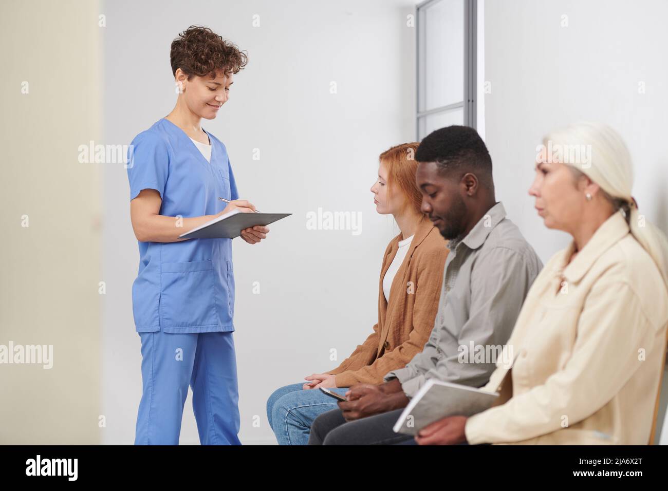 Gruppo di tre persone multietniche in coda in attesa di consultazione in ospedale moderno, operatore medico in piedi vicino alla presenza di controllo Foto Stock