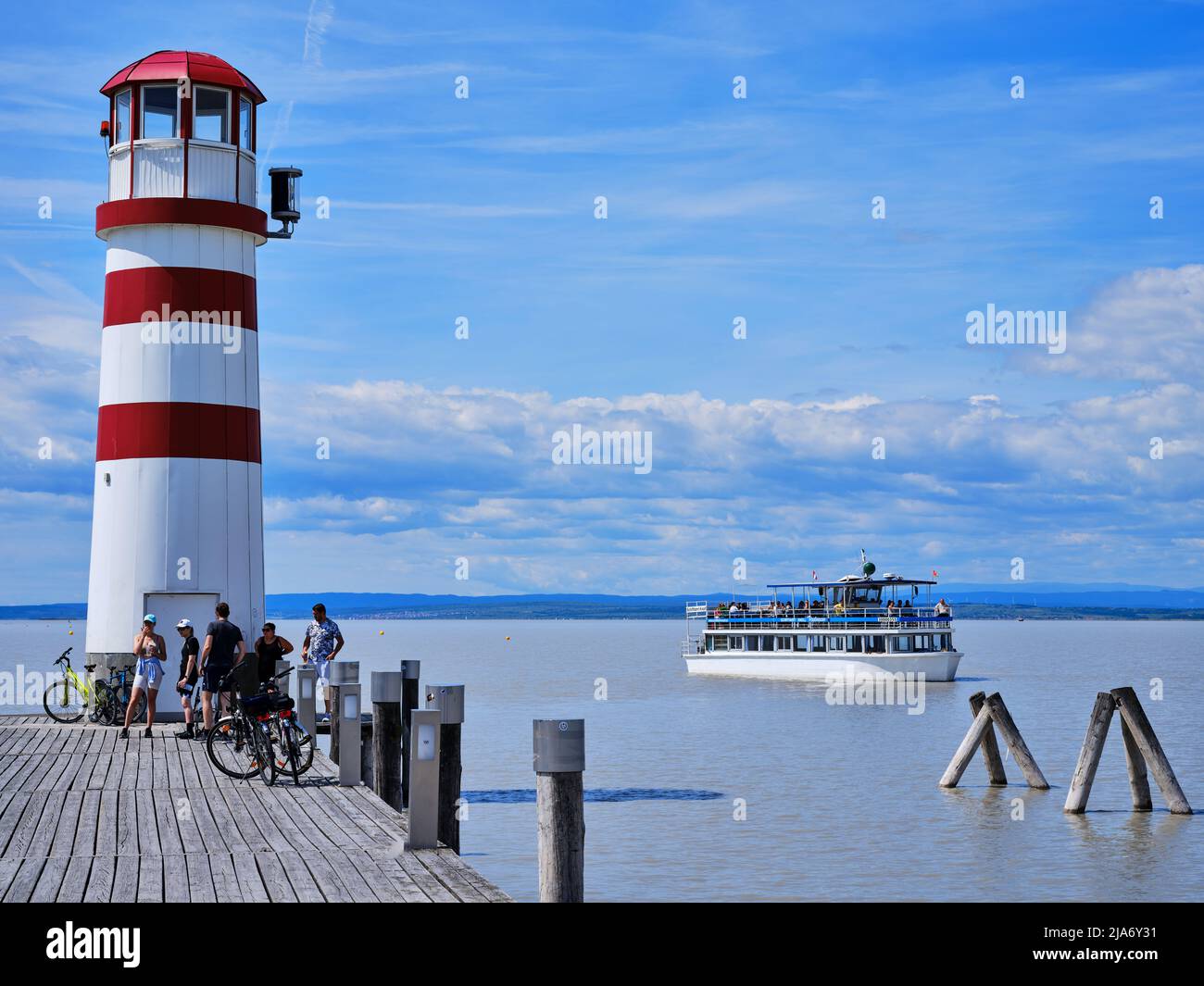 Faro rosso e bianco a Podersdorf am SEE, Austria, con il lago Neusiedl, traghetto e ciclisti su un molo di legno. Foto Stock