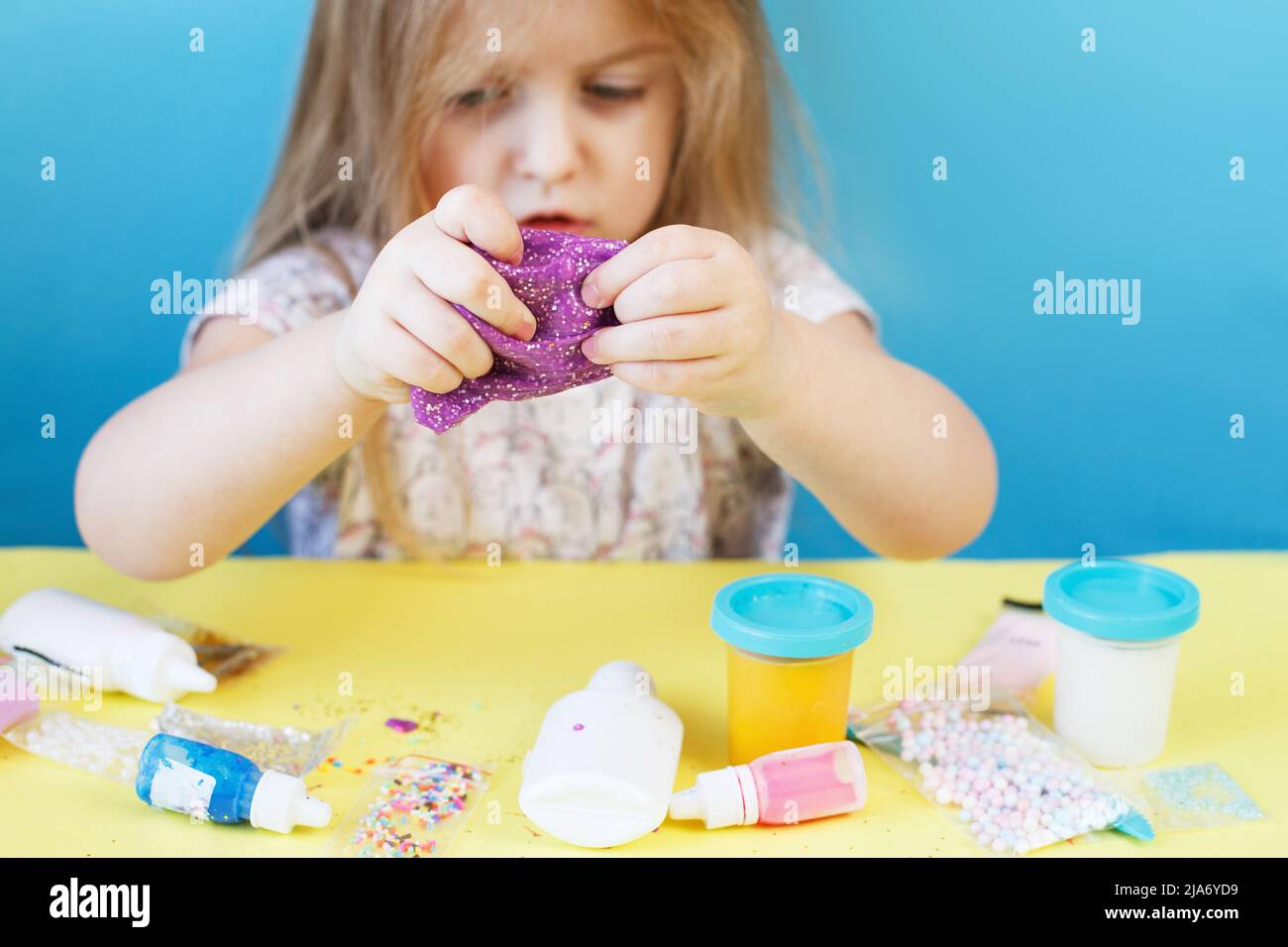 La ragazza bionda tiene il glime viola isolato su uno sfondo blu. Bambino che gioca con un giocattolo lime. Fare slime. Spazio di copia. Concetto DIY. Foto Stock