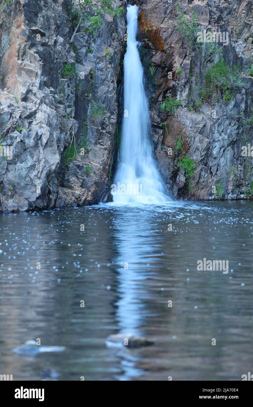 Alta cascata che scorre tra pareti di roccia verticali nella Sierra de Madrid, in Spagna, con una tranquilla piscina riflettente alla base e vegetazione naturale di montagna Foto Stock