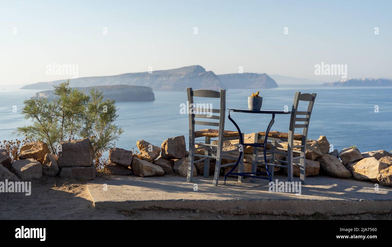 Un posto con vista: Un tavolo singolo con due sedie occupa un posto con una vista mozzafiato della caldera di Santorini nella luce del tardo pomeriggio Foto Stock