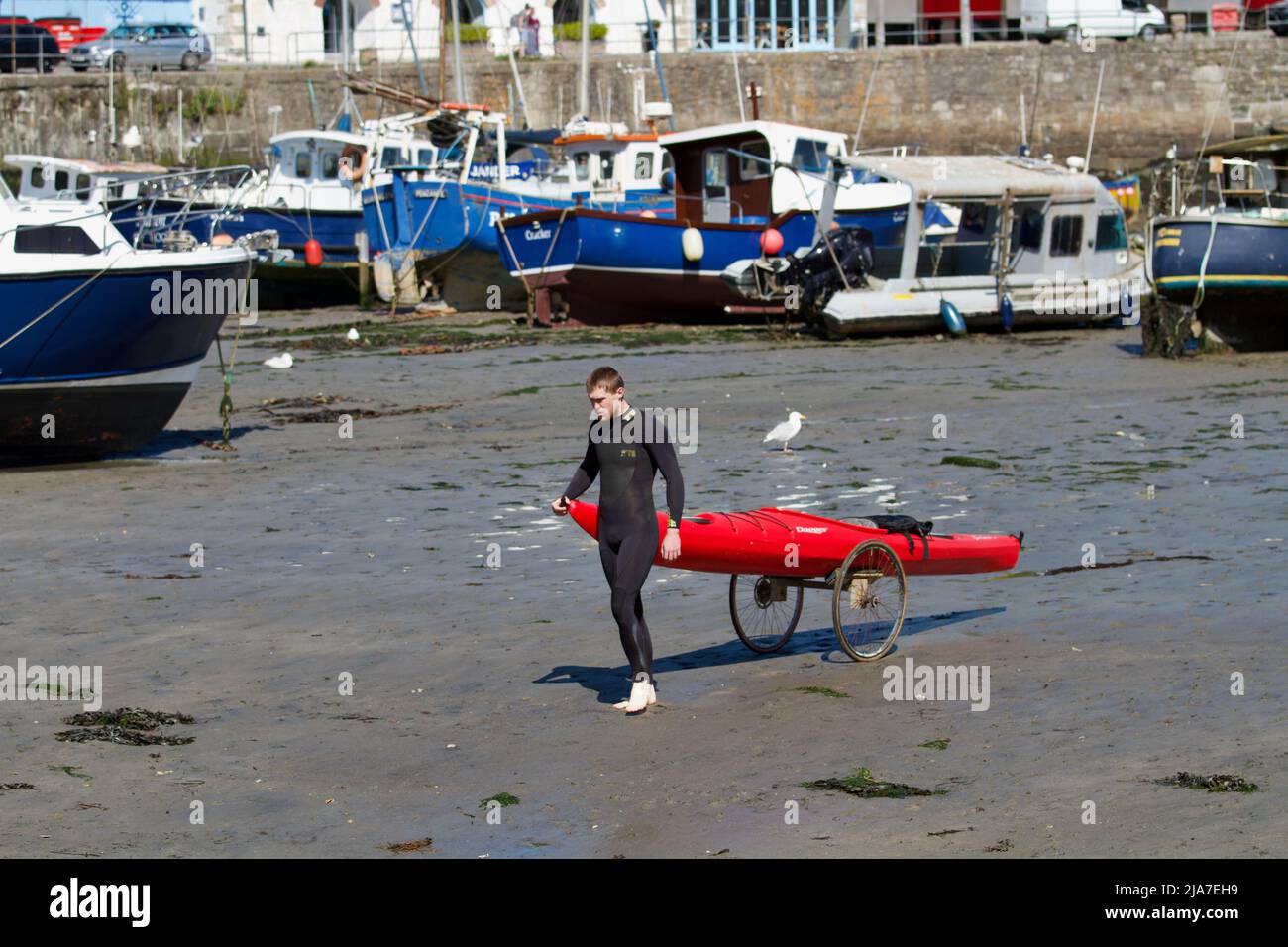 Giovane uomo in muta con il suo kayak su un trolly nel porto di Porthleven a bassa marea Foto Stock